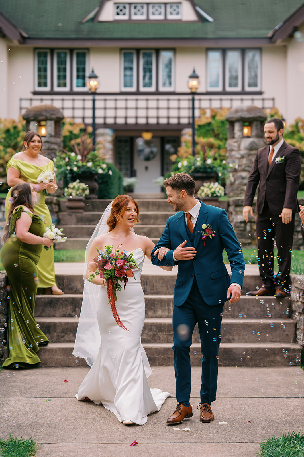 Bride and groom wearing walking down mansion steps with green bridesmaids and brown suit groomsmen