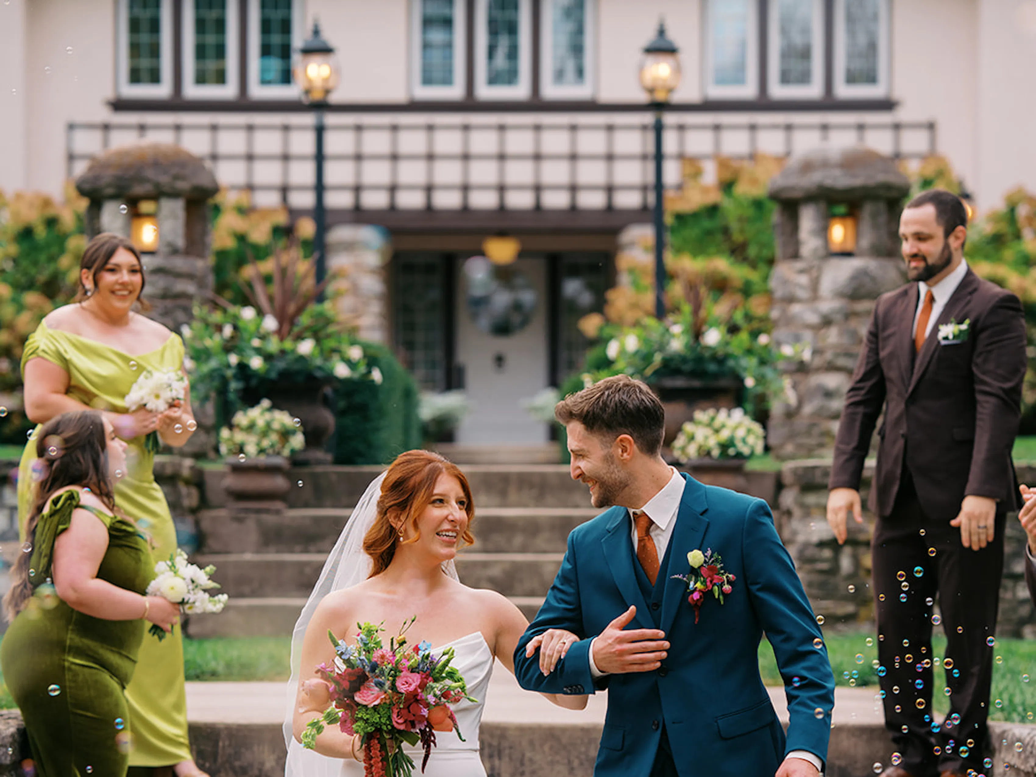 Bride and groom wearing walking down mansion steps with green bridesmaids and brown suit groomsmen