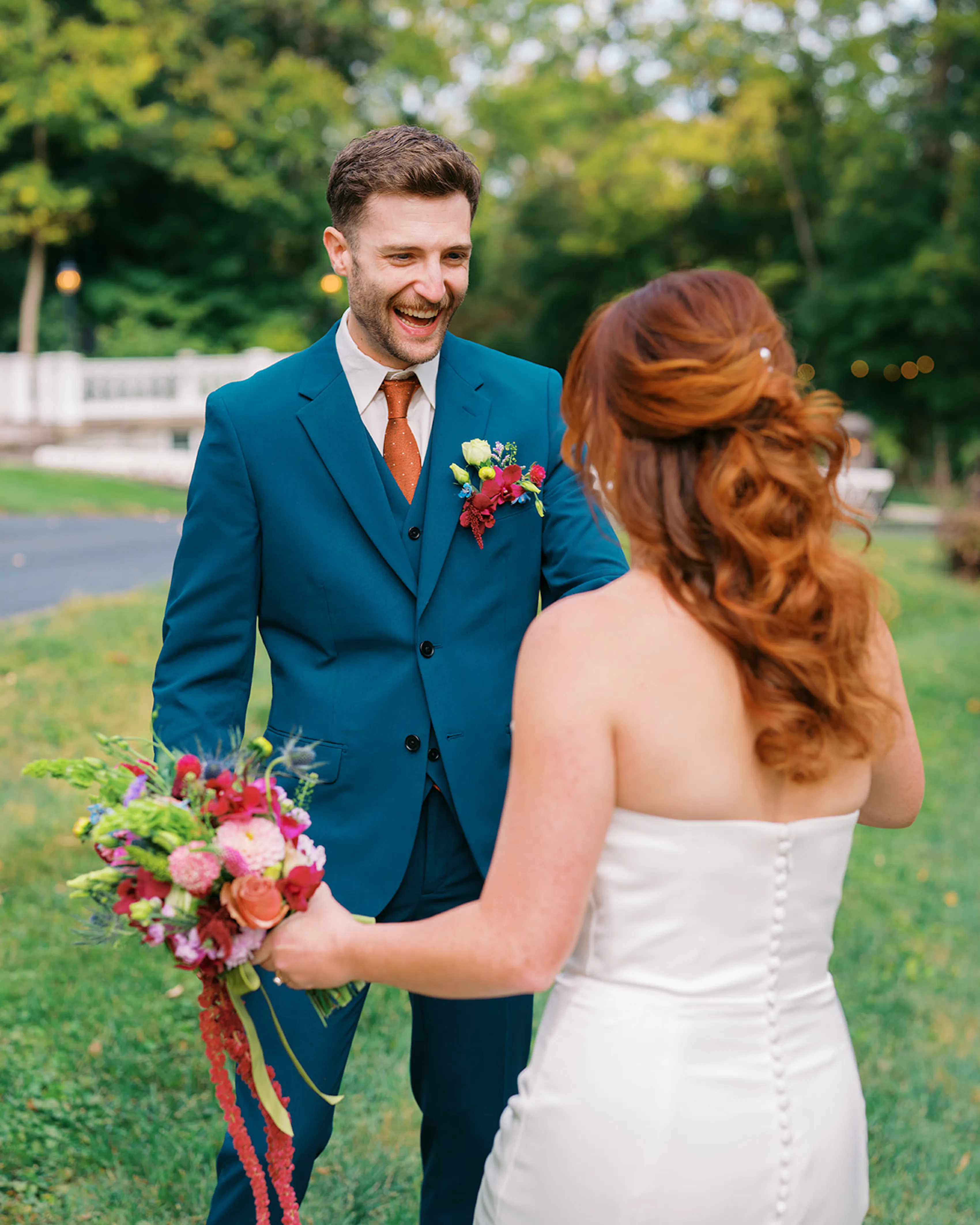 First look with excited groom in dark teal suit and rust tie with redhead bride carrying colorful bouquet