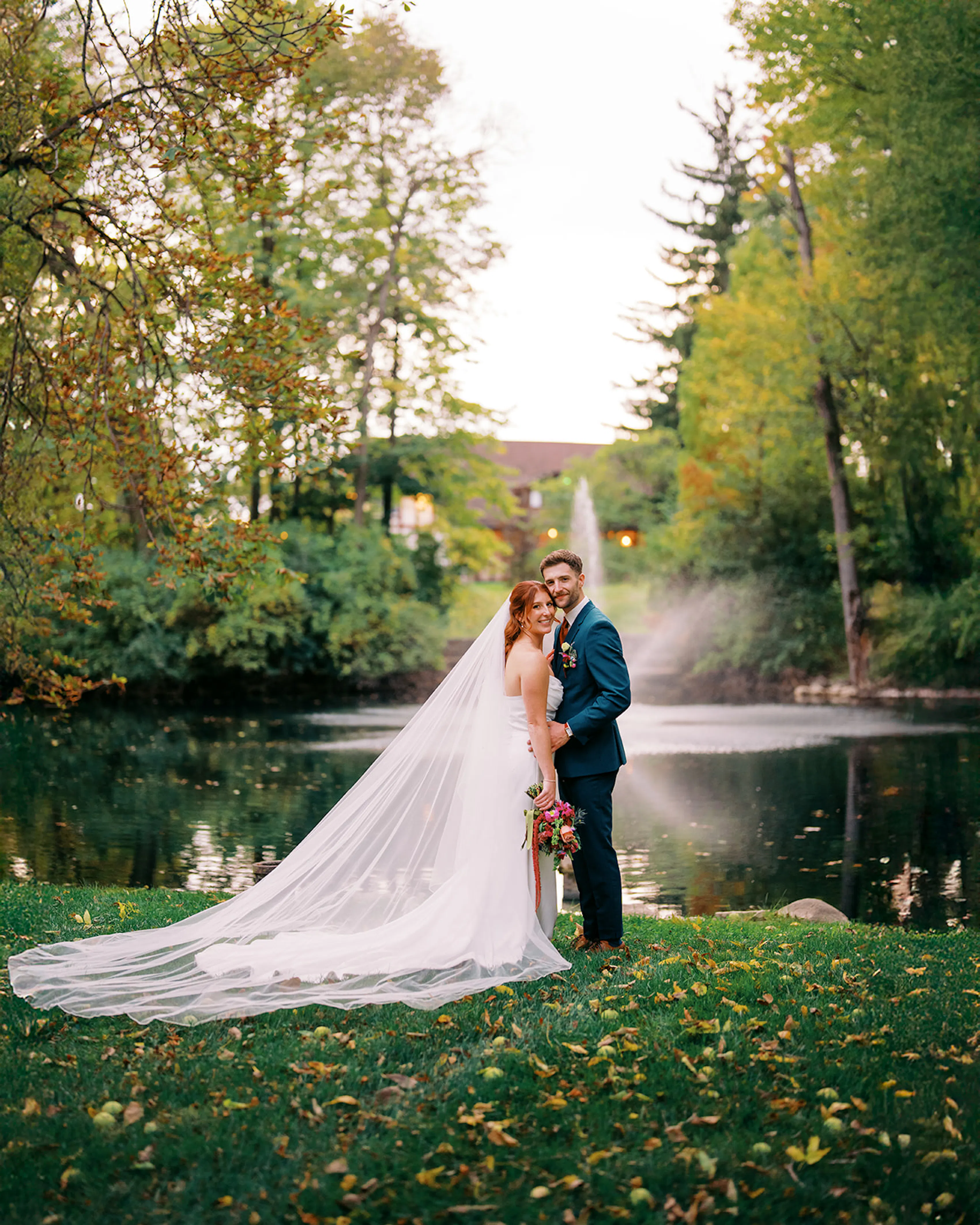 Bride and groom in front of estate water feature in the fall wearing veil and teal suit