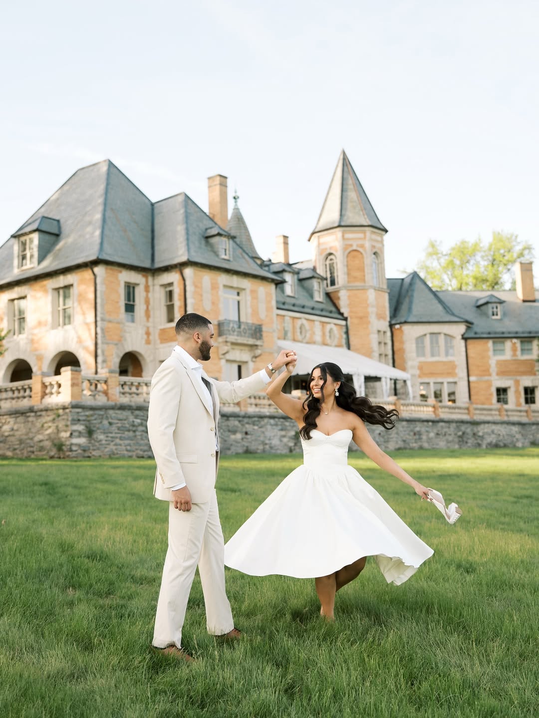 Bride in midi gown and groom in tan suit dancing outside estate wedding venue