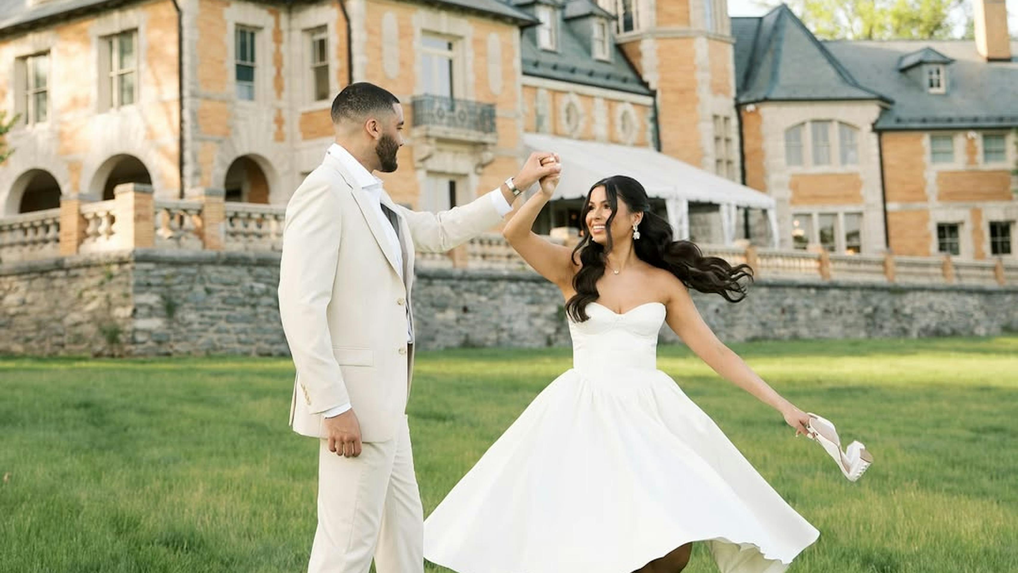 Bride in midi gown and groom in tan suit dancing outside estate wedding venue