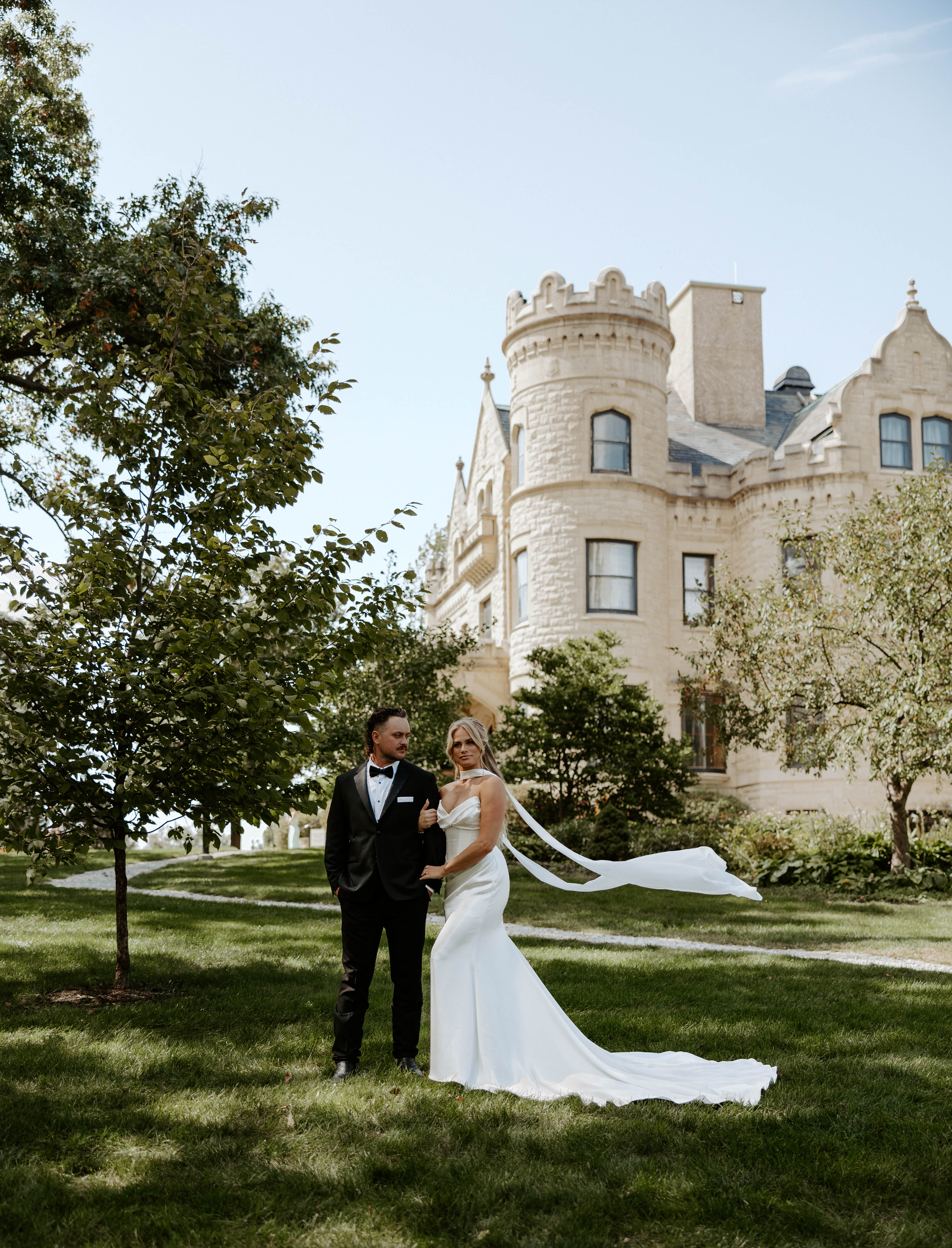 Summer wedding at castle venue with neck scarf wedding gown and black groom tuxedo