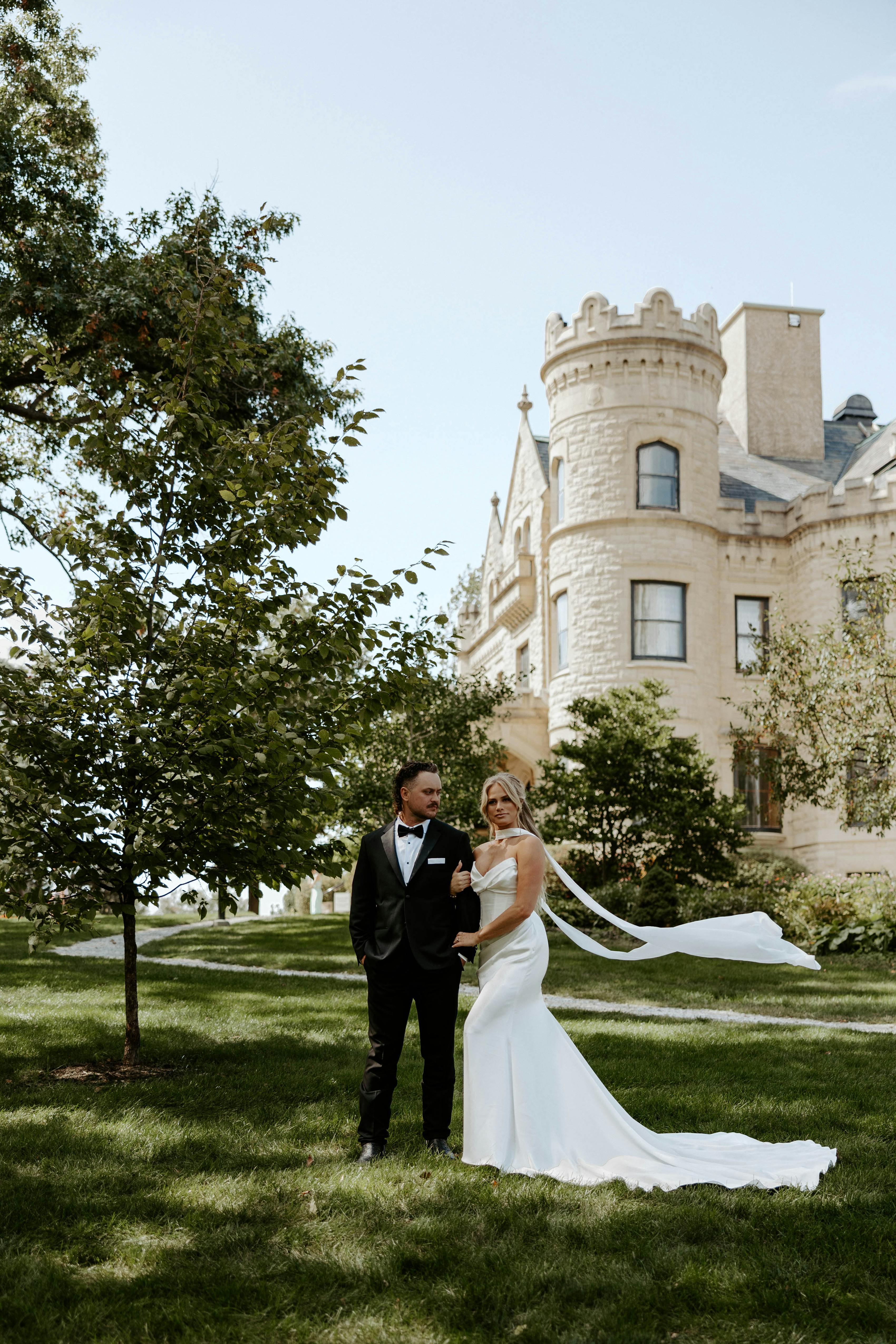 Summer wedding at castle venue with neck scarf wedding gown and black groom tuxedo