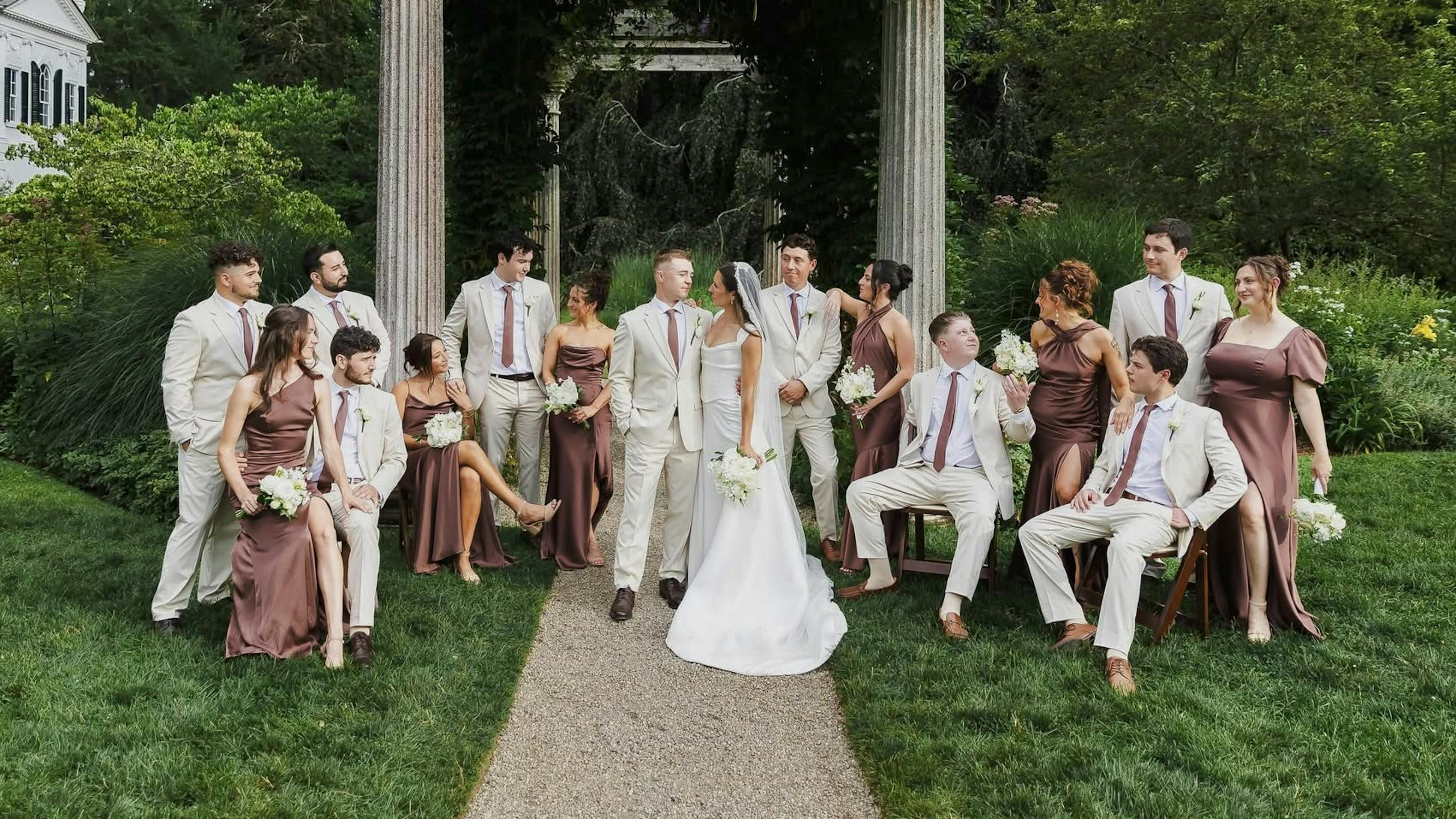 Outdoor wedding with tan groomsmen suits and coordinating dark brown satin bridesmaid dresses.