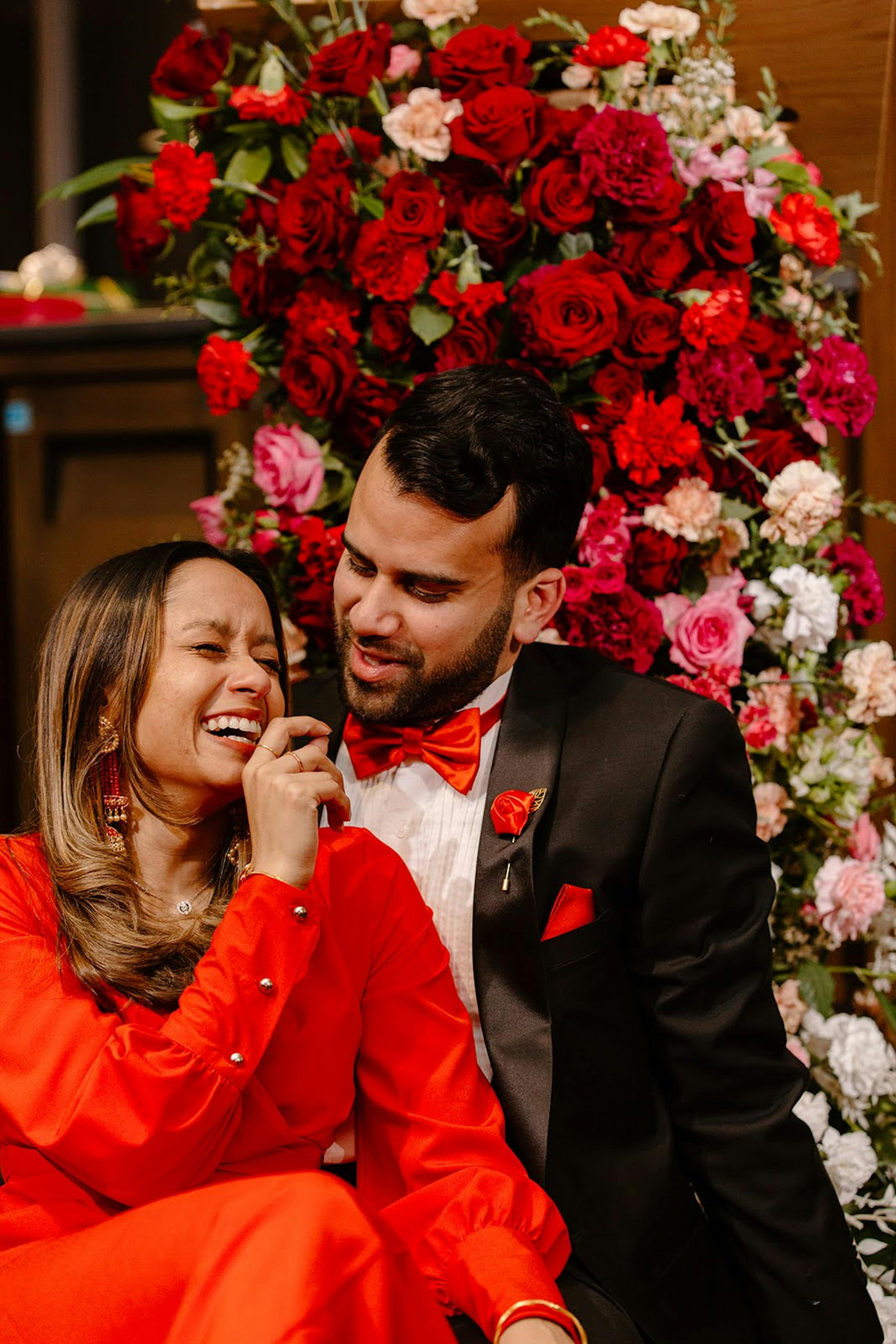 Valentine's Day engagement wearing red dress and black tuxedo with red bow tie in front of flower backdrop