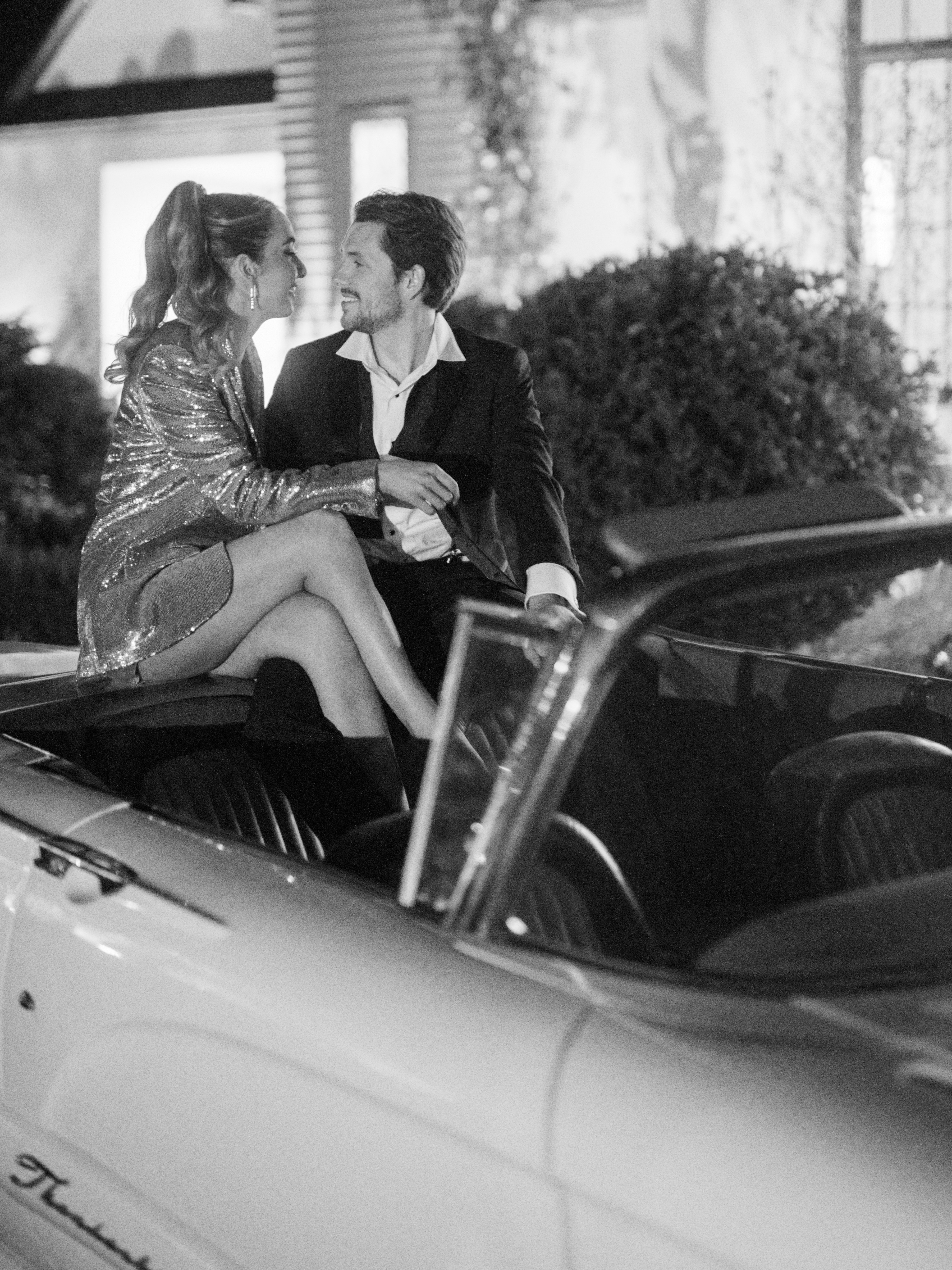 Romantic couple wearing dress and suit sitting on parked vintage convertible car