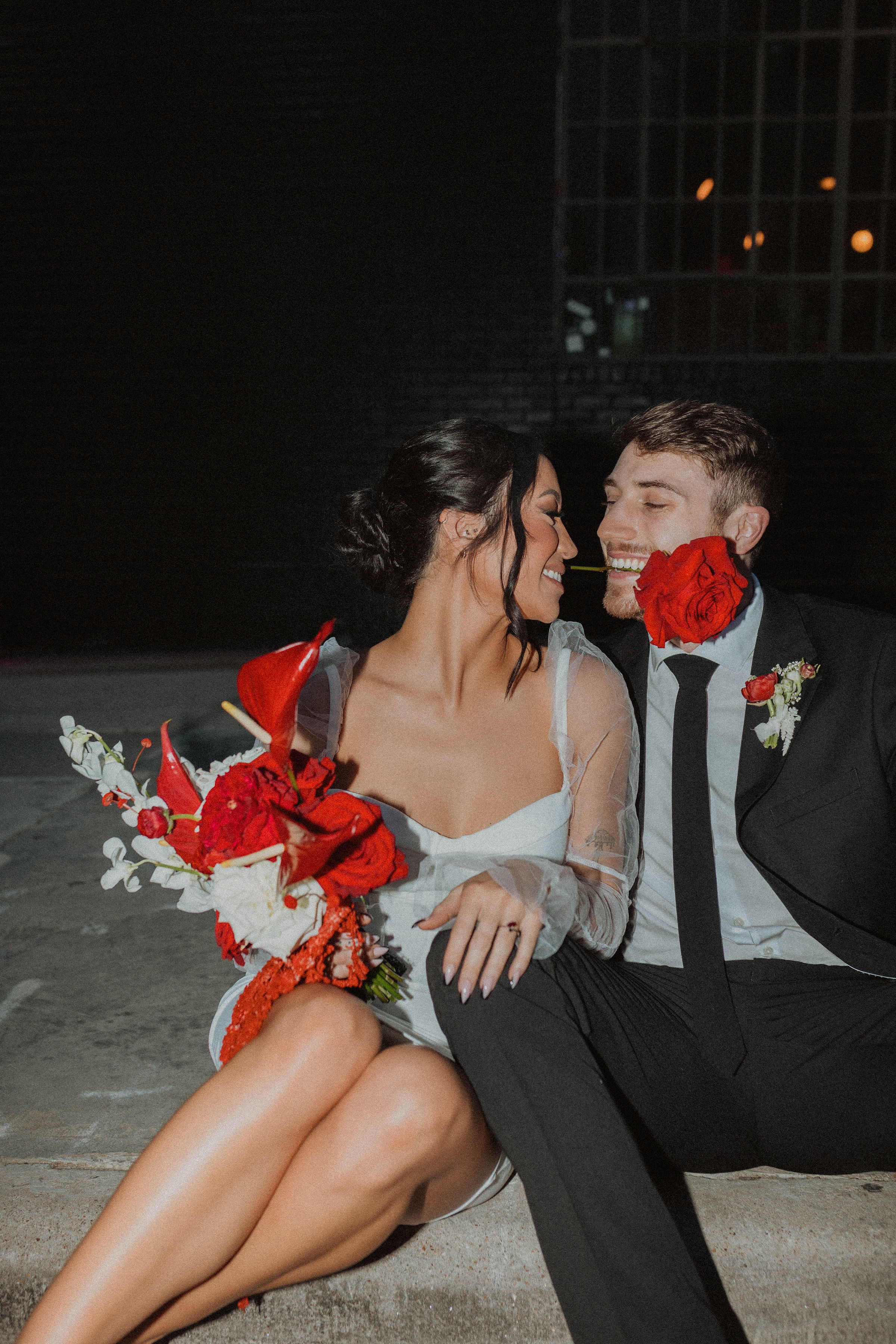 Couple wearing white short dress and black suit, holding red flowers, posed with rose in mouth