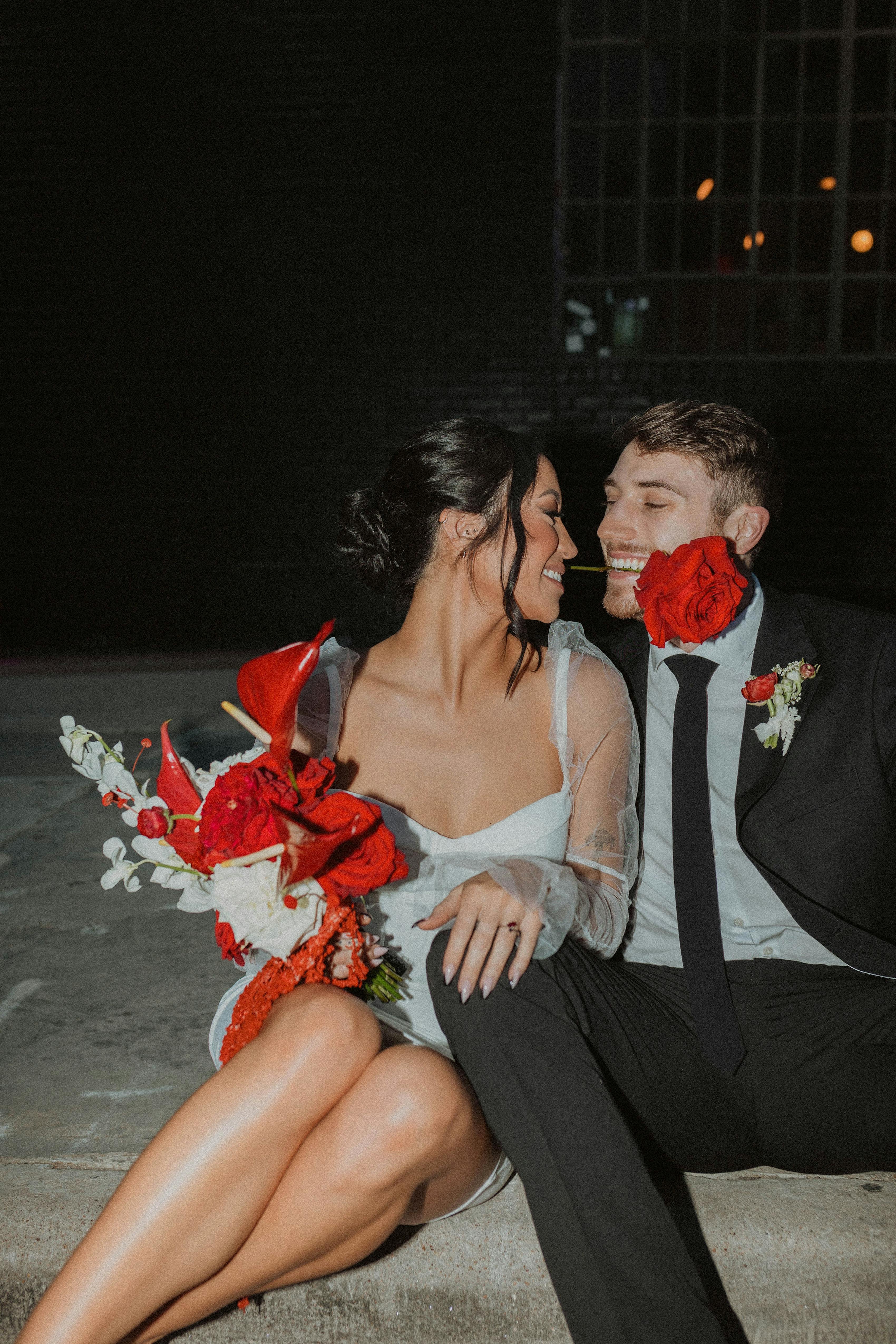Couple wearing white short dress and black suit, holding red flowers, posed with rose in mouth