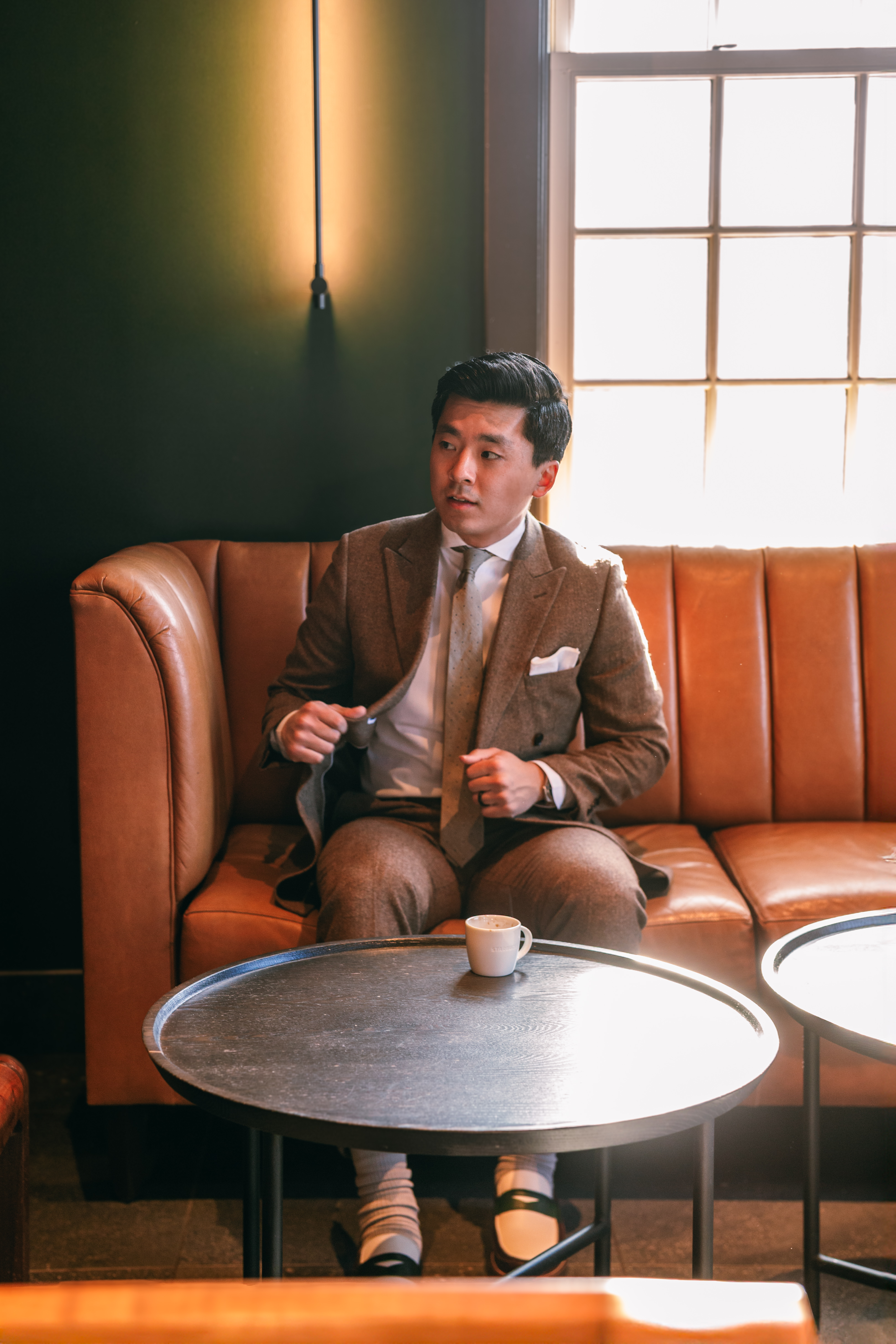 Man wearing brown tweed suit sitting on couch at cafe drinking coffee