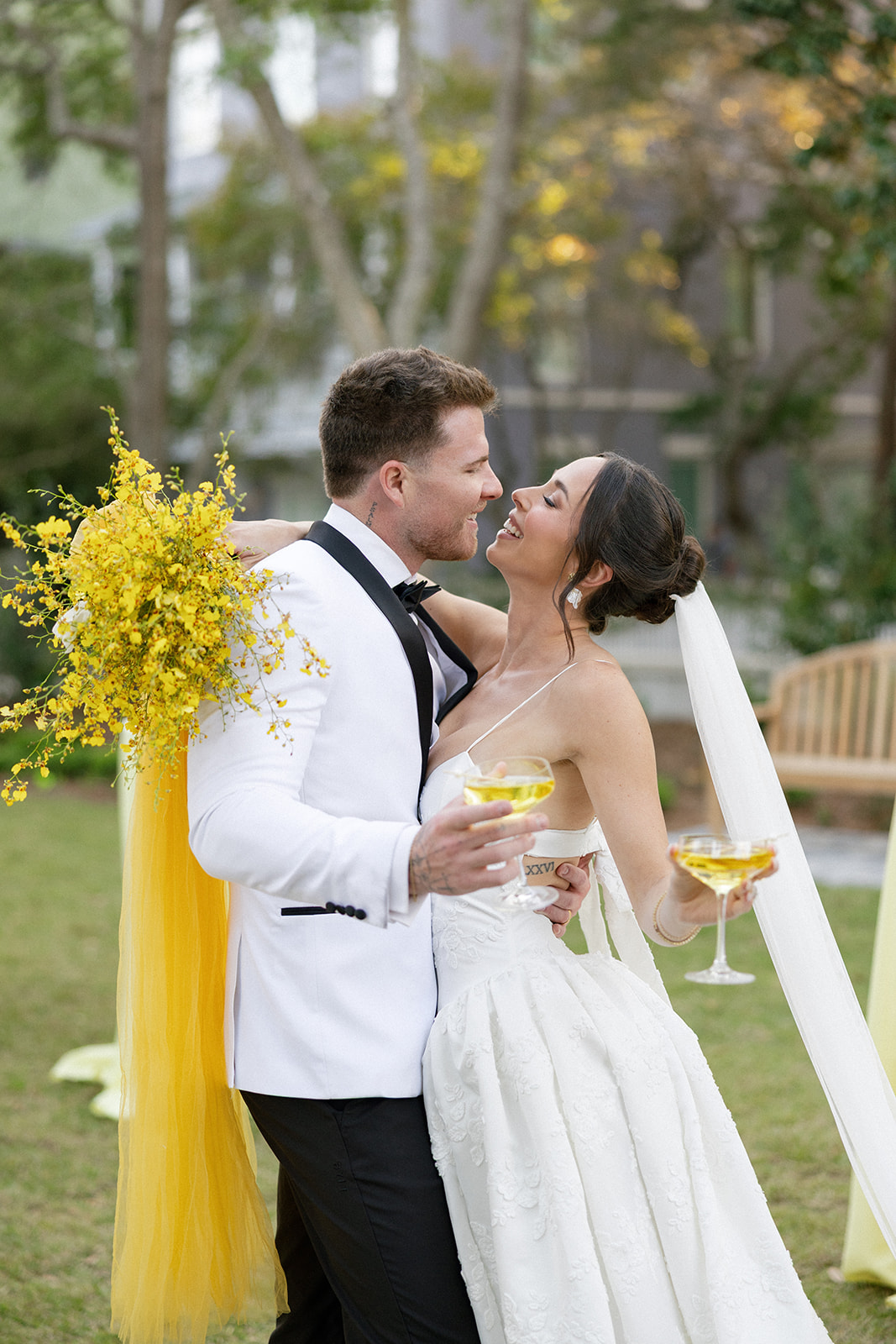 Groom wearing ivory tux jacket with black lapels kissing bride holding yellow bouquet and tulle