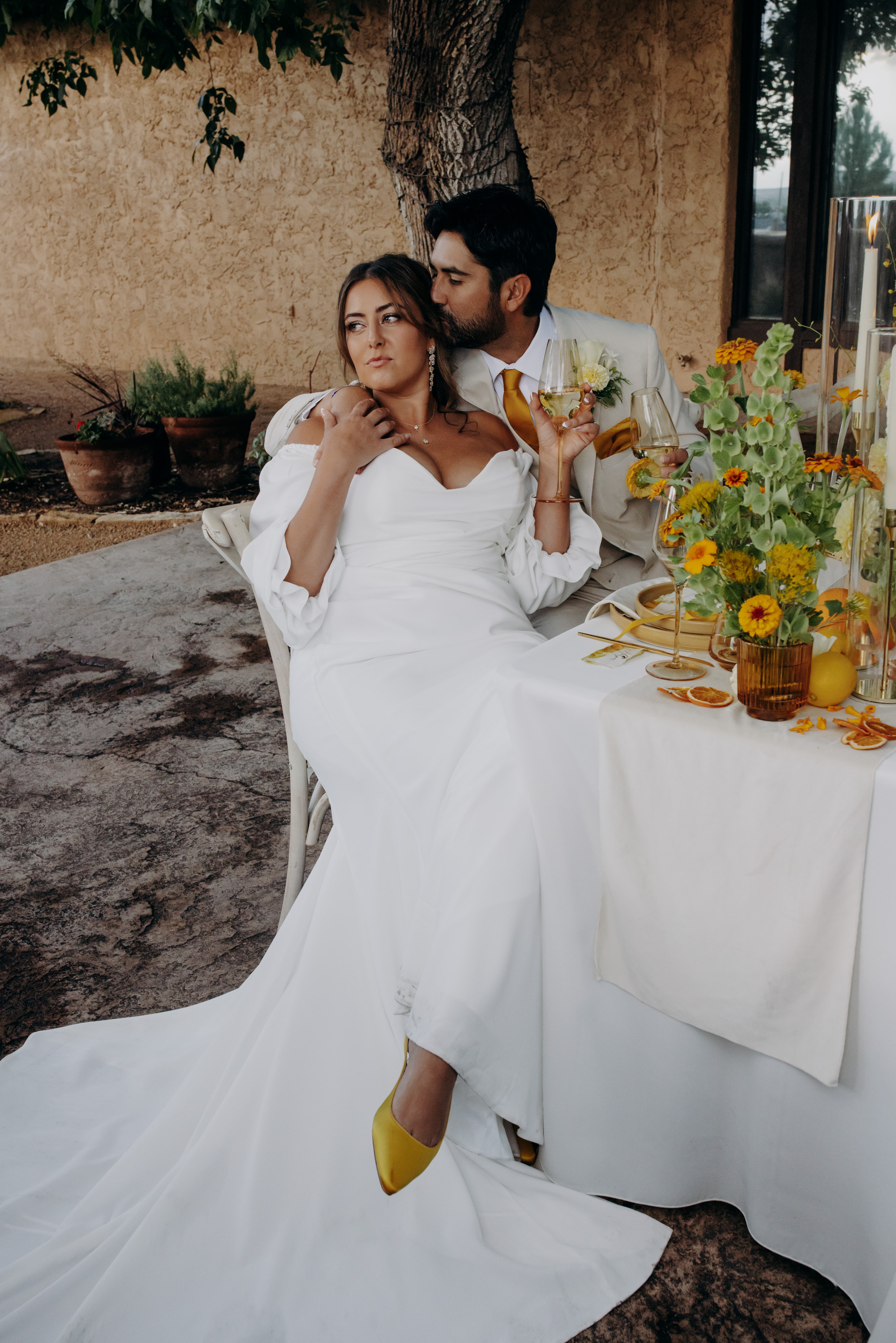 Bride wearing yellow shoes sitting with groom wearing tan suit and orange tie next to bright floral tablescape