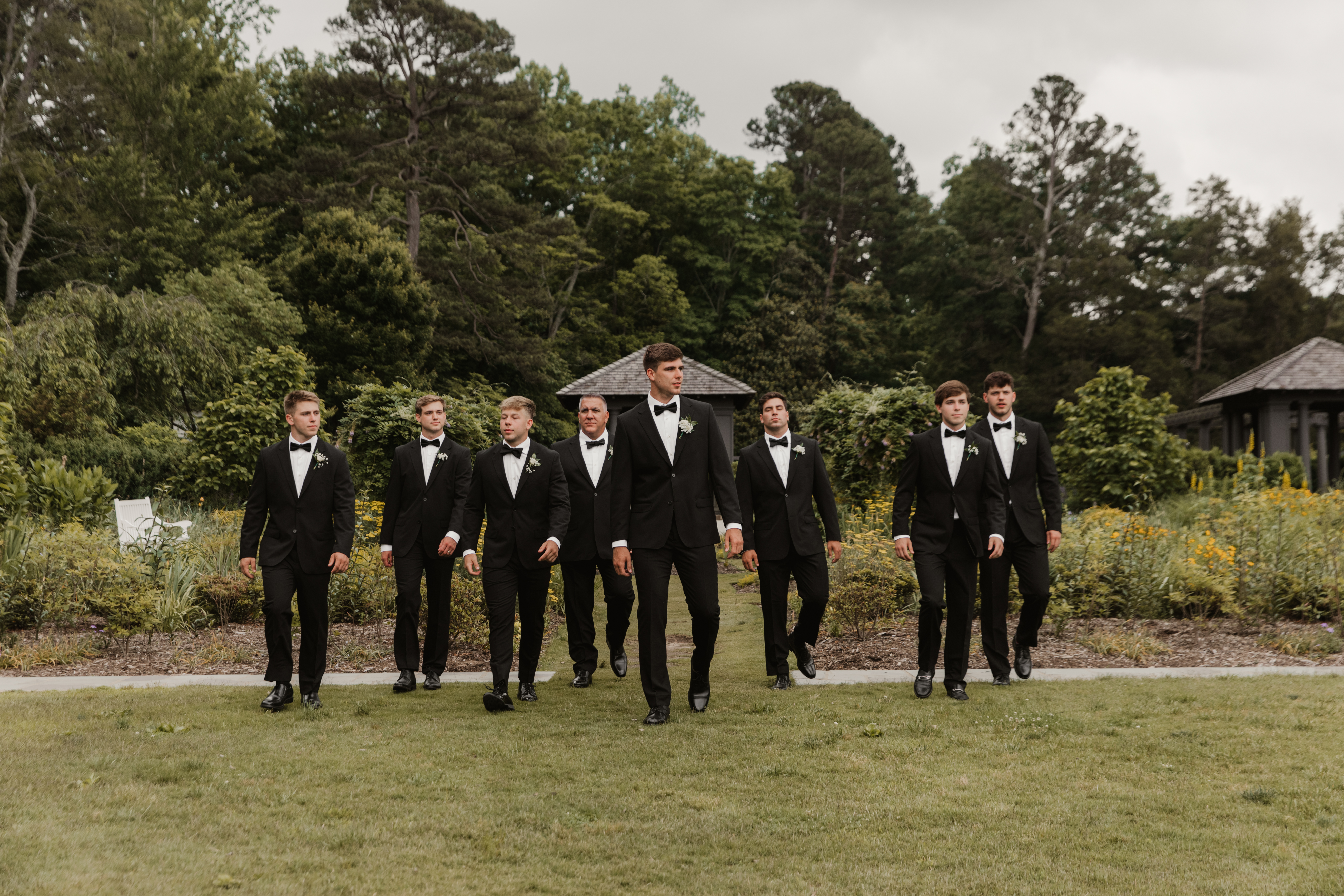 A groom and his groomsmen in black tuxedos walking as a group outside on a patch of grass.