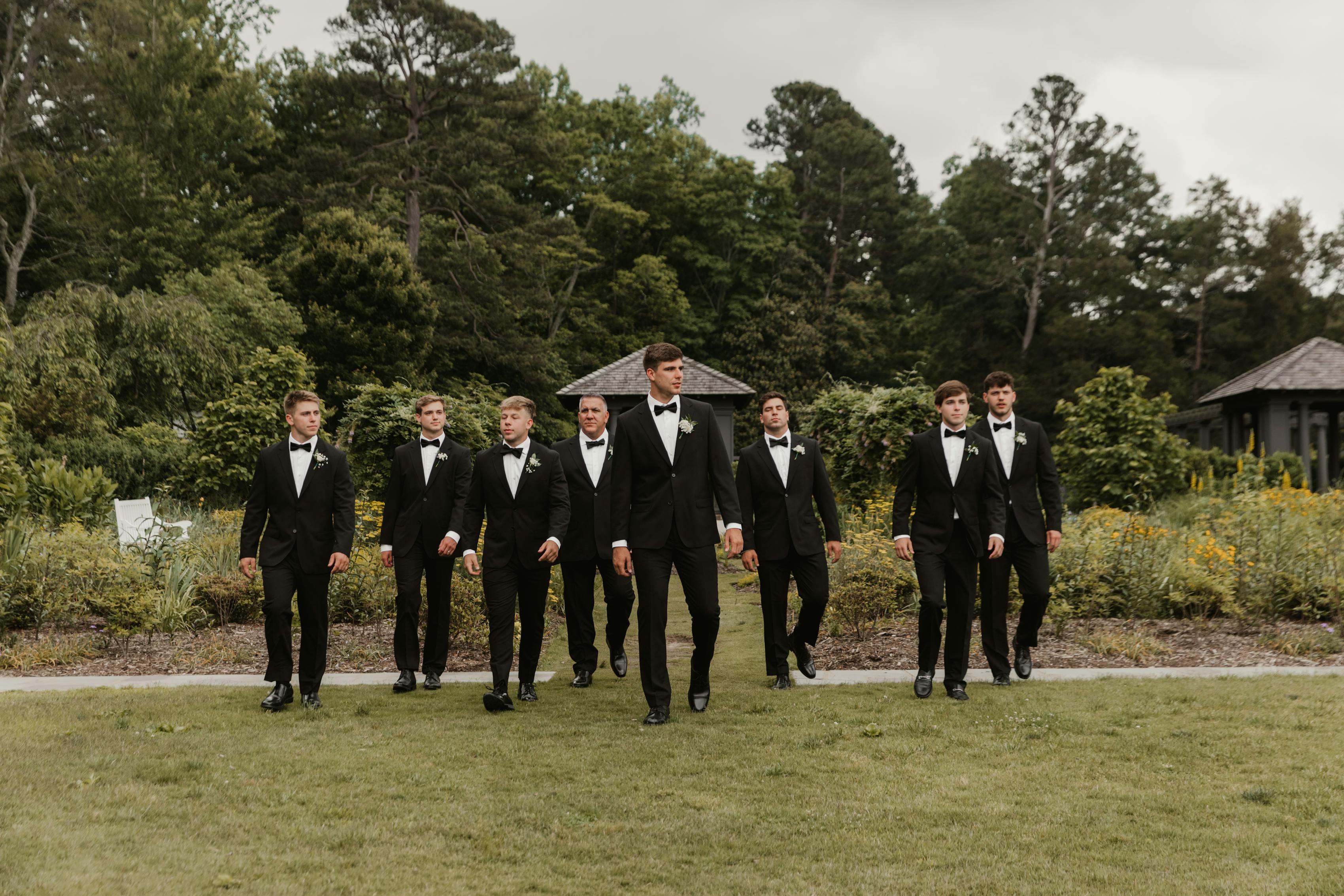 A groom and his groomsmen in black tuxedos walking as a group outside on a patch of grass.