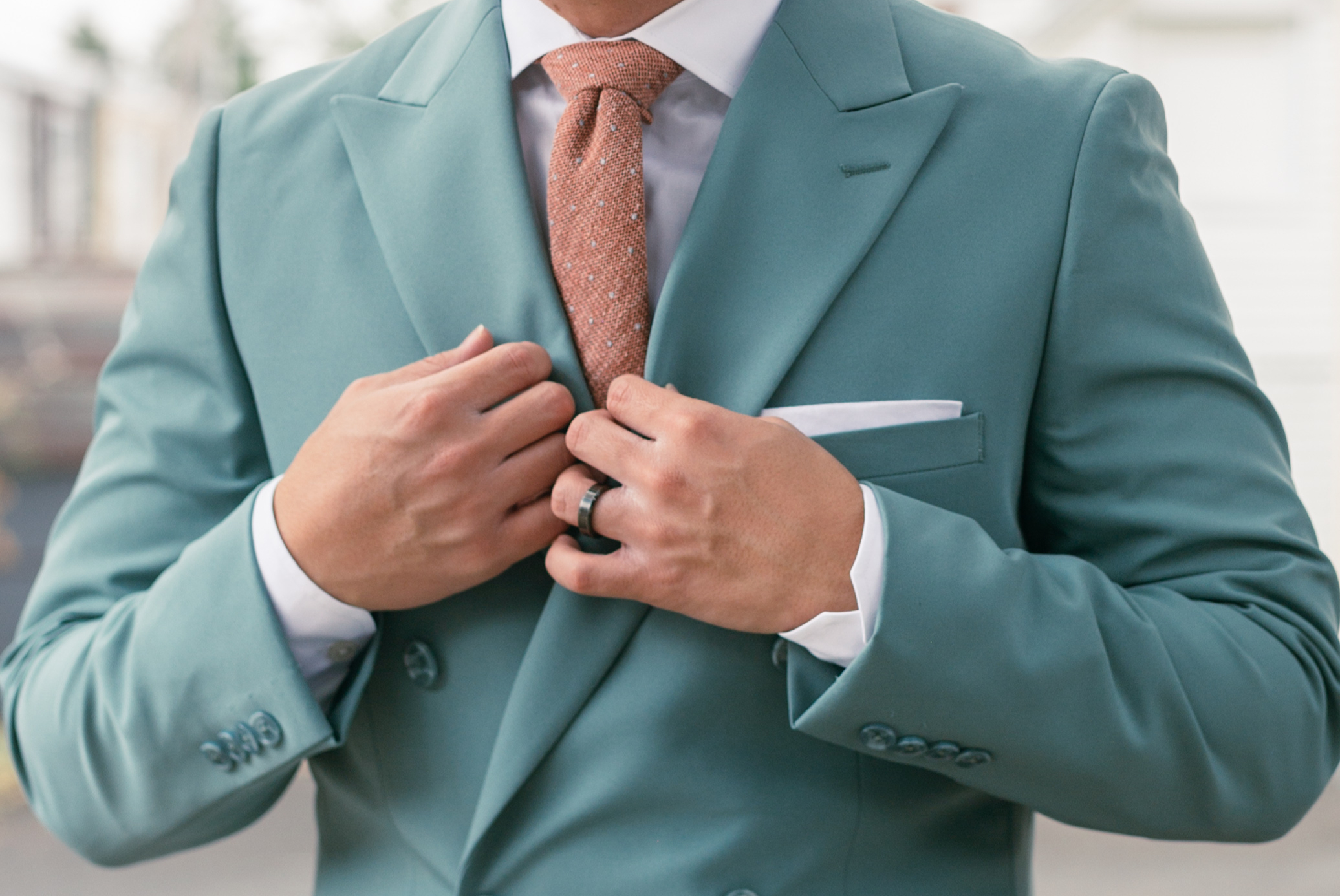 Close-up image of a man in a blue suit with a salmon colored tie with his hands on his suit, slightly covering his white pocket square.
