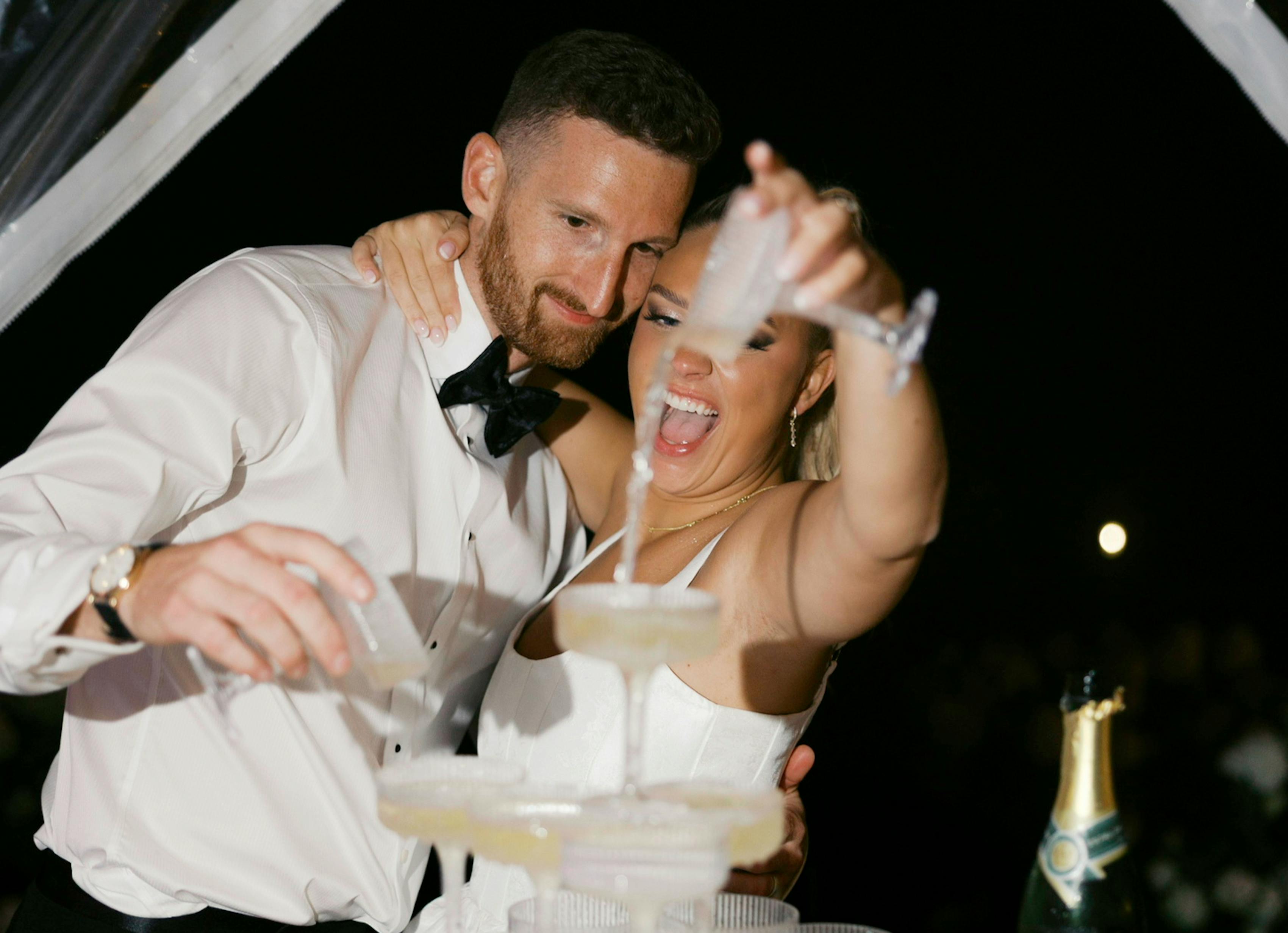 Newlywed couple holding eachother while pouring champagne into a tower of glasses at night.