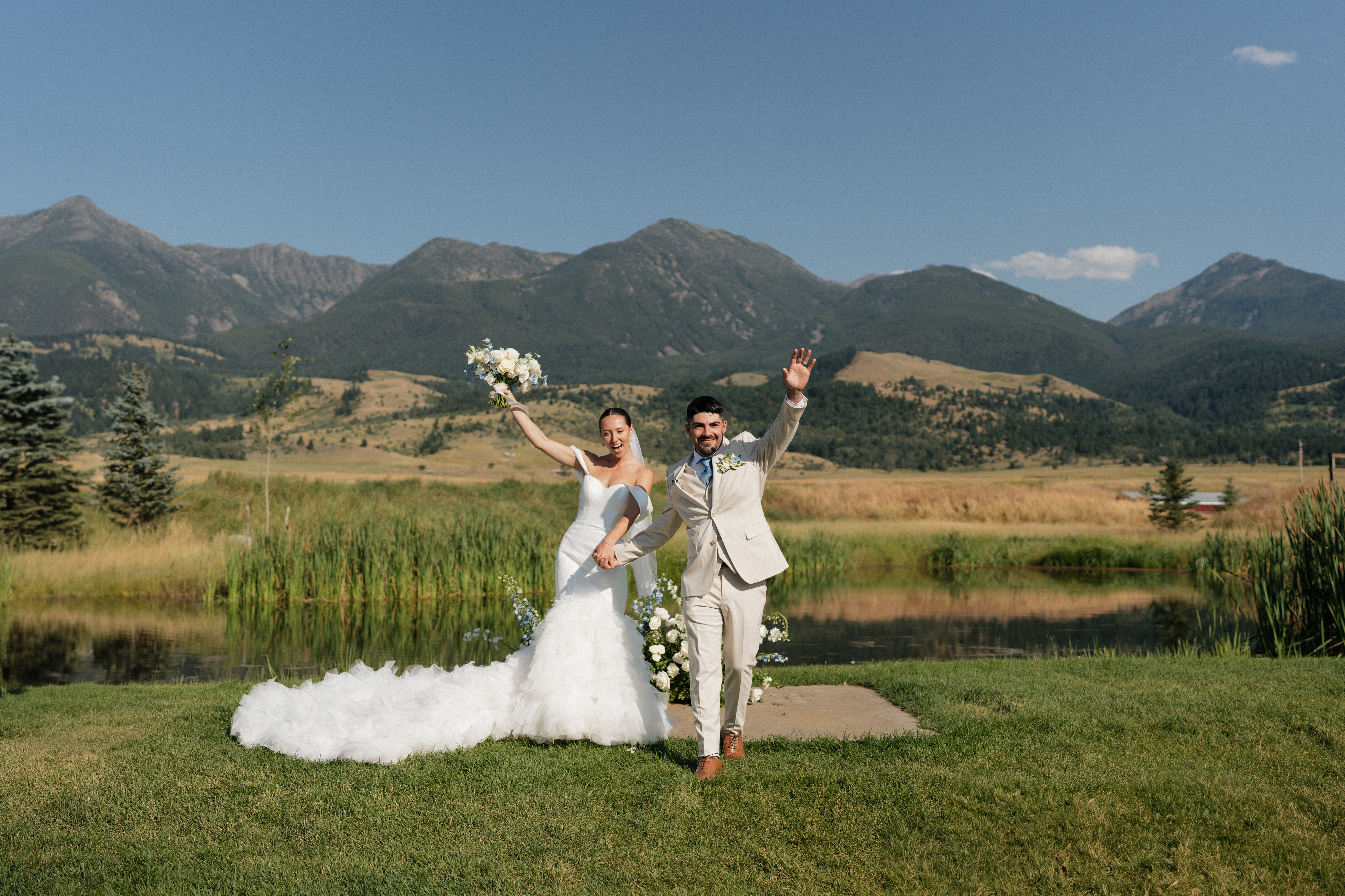 A bride in a feathered white gown and a groom in a tan suit celebrate their new marriage in front of beautiful mountains over a lake.