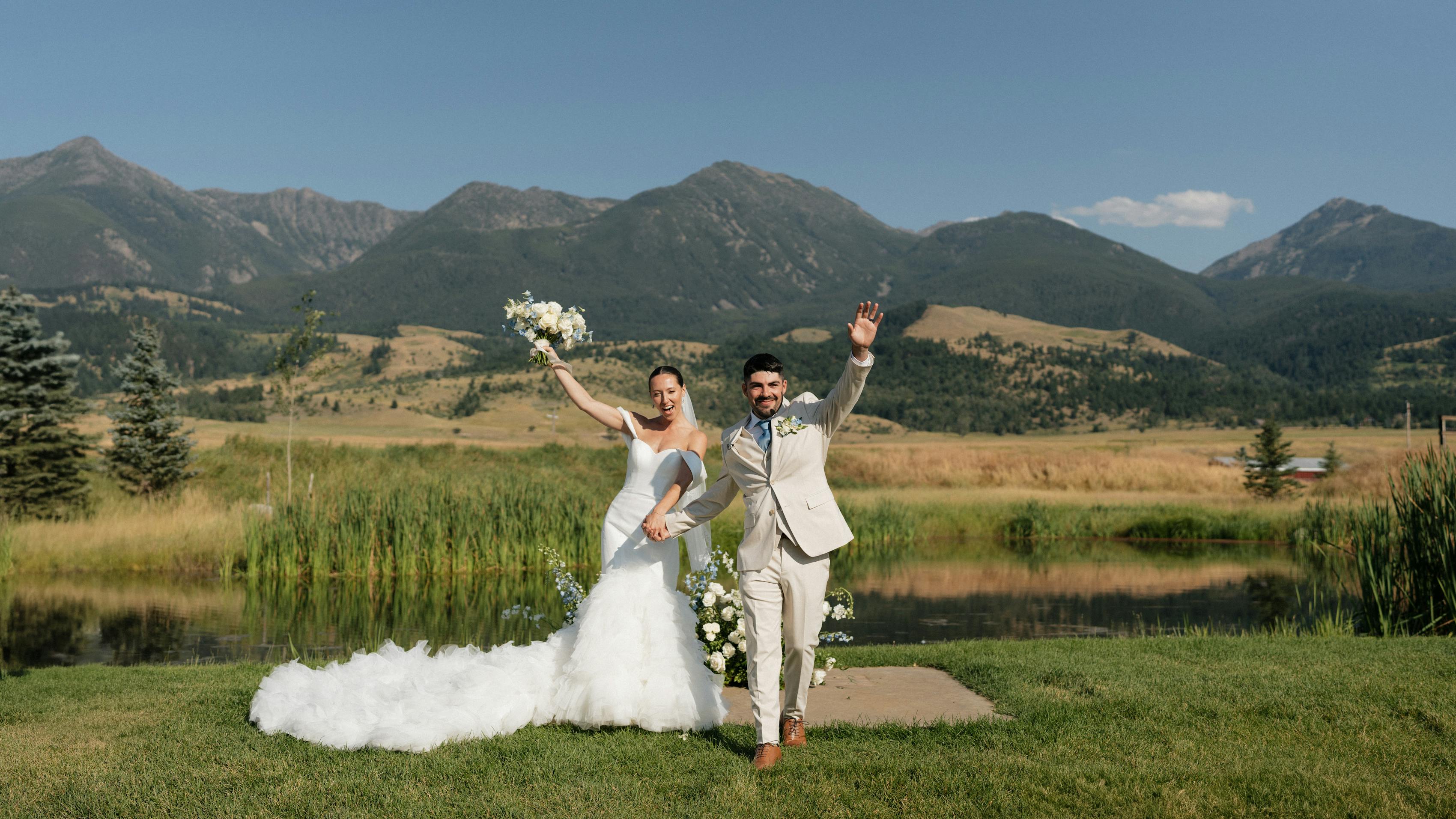 A bride in a feathered white gown and a groom in a tan suit celebrate their new marriage in front of beautiful mountains over a lake.