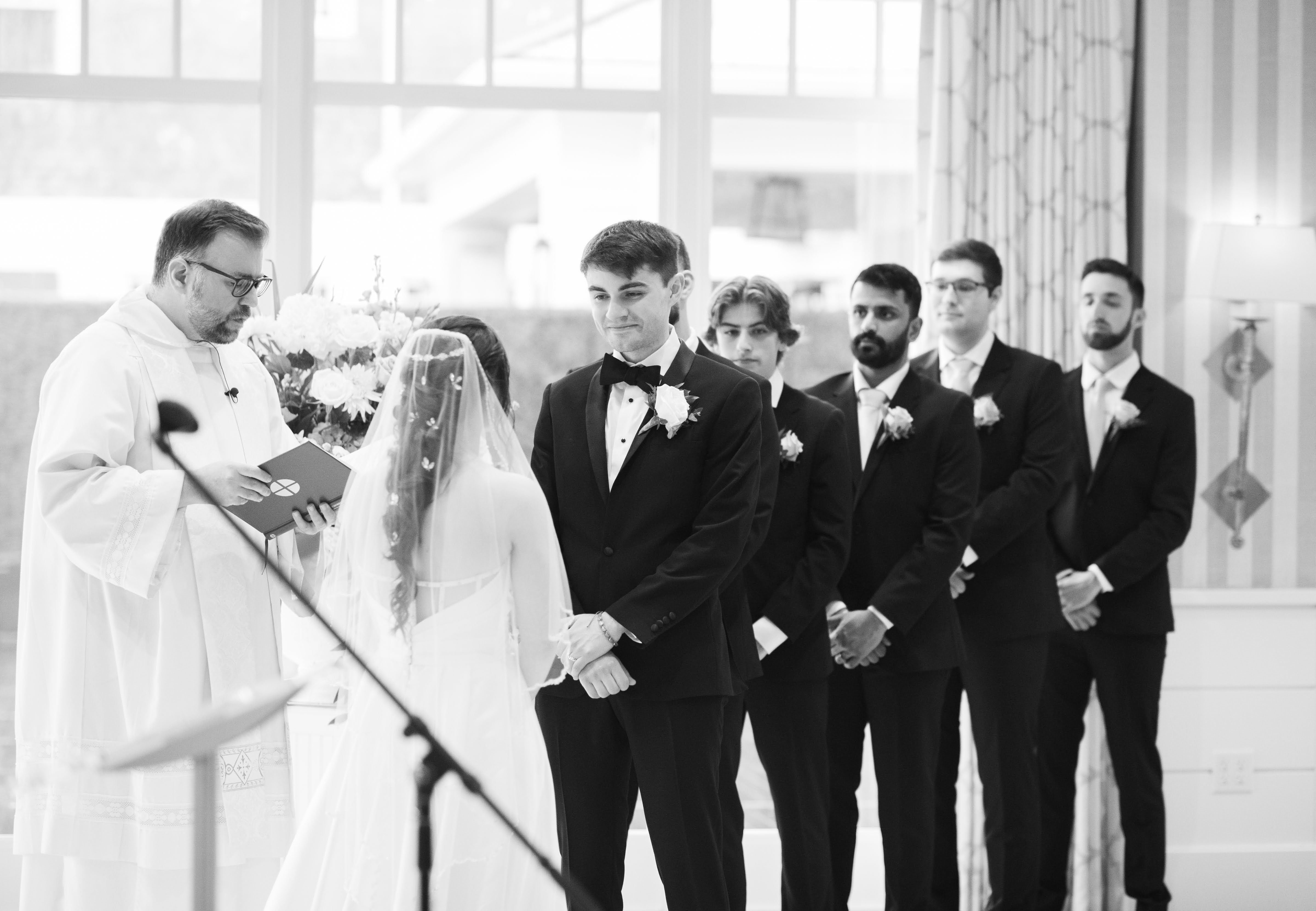Black and white photo of an ordained minister wedding a bride and groom. From this angle, we can see the back of the bride, the front of the groom, and all of the groomsmen.