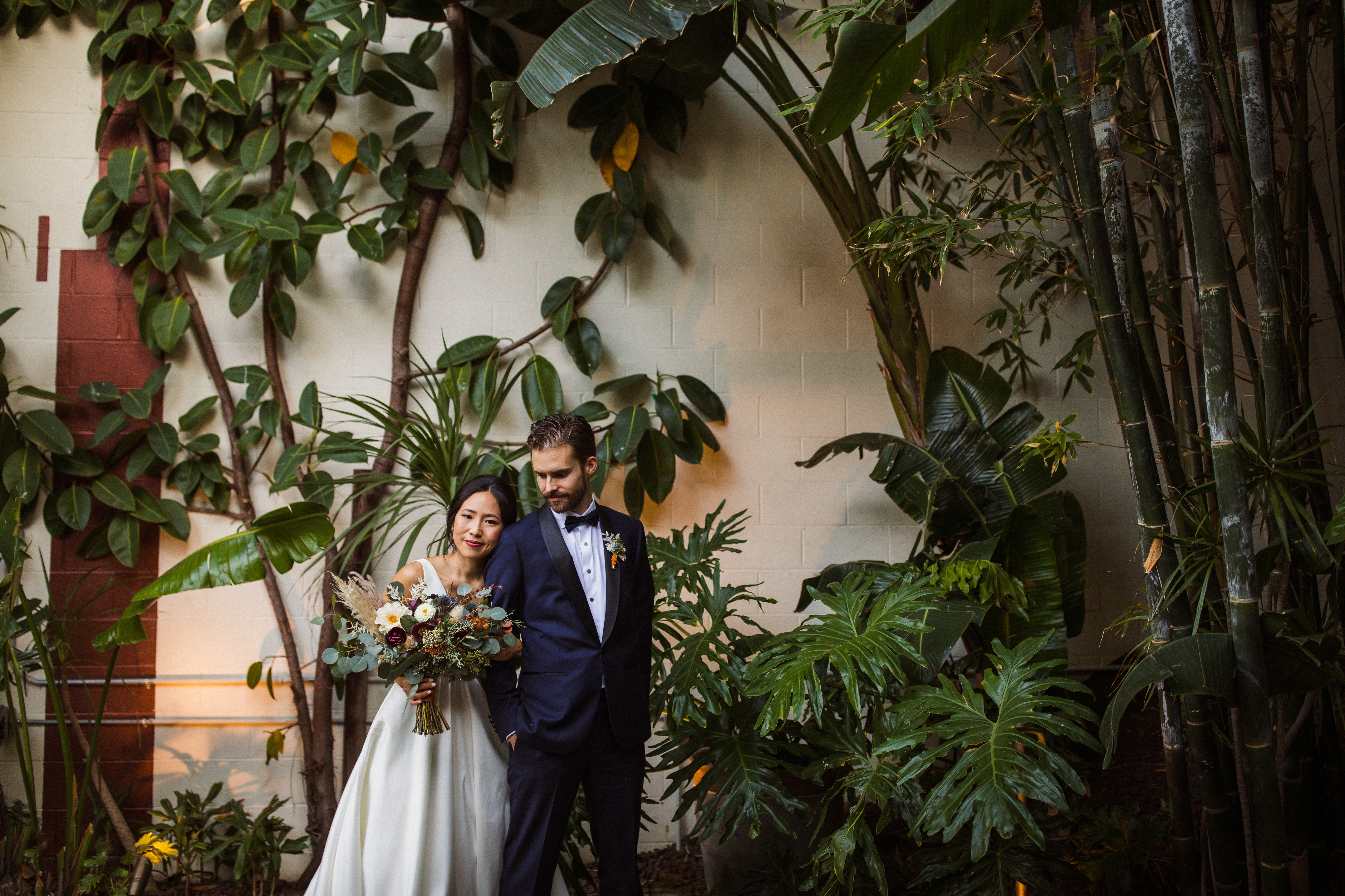 Bride and groom in A-line dress and navy tuxedo with modern tropical decor greenery installation