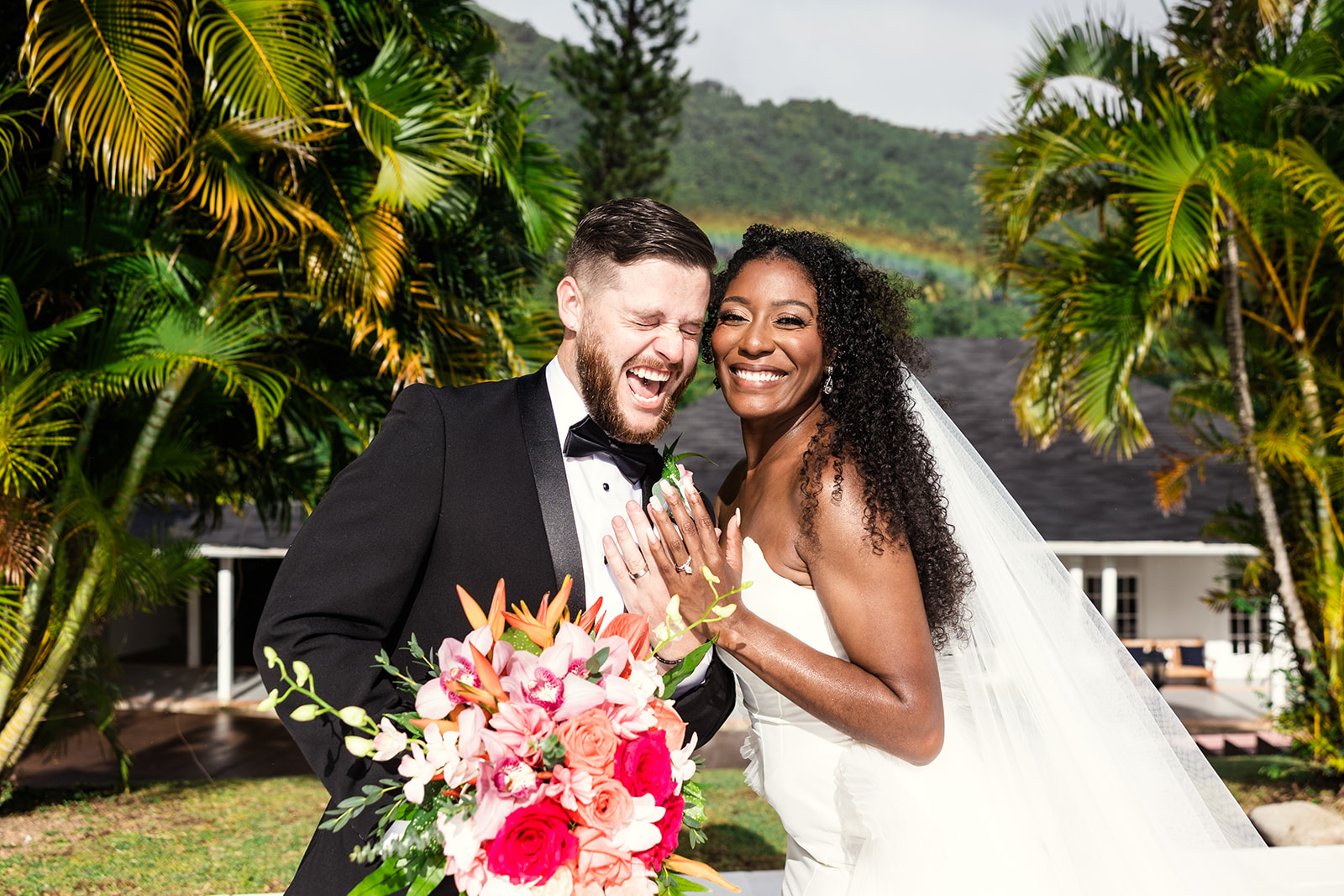 Black tie bride and groom beaming in front of rainbow and palm trees at tropical wedding