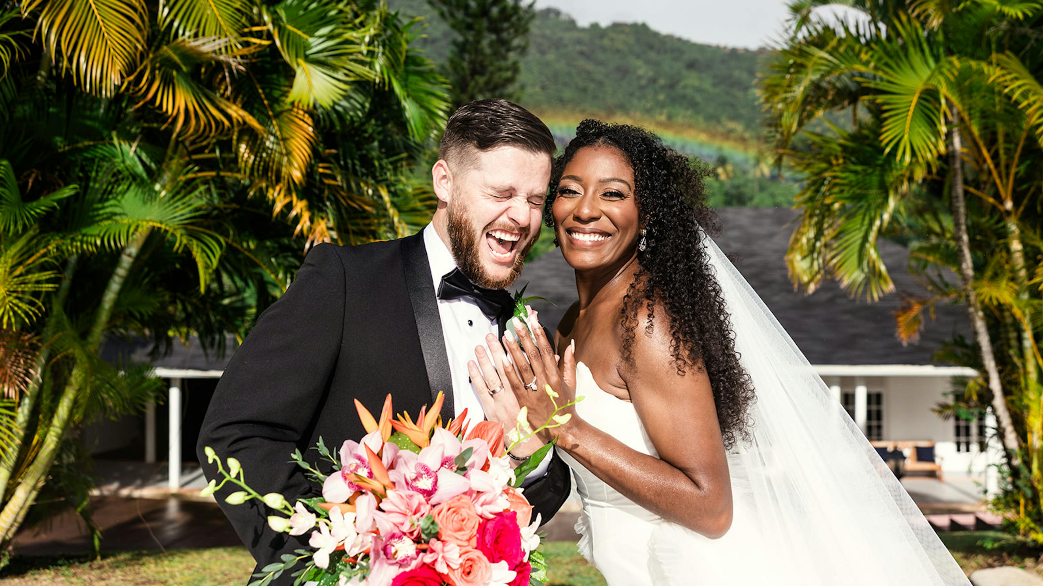 Black tie bride and groom beaming in front of rainbow and palm trees at tropical wedding