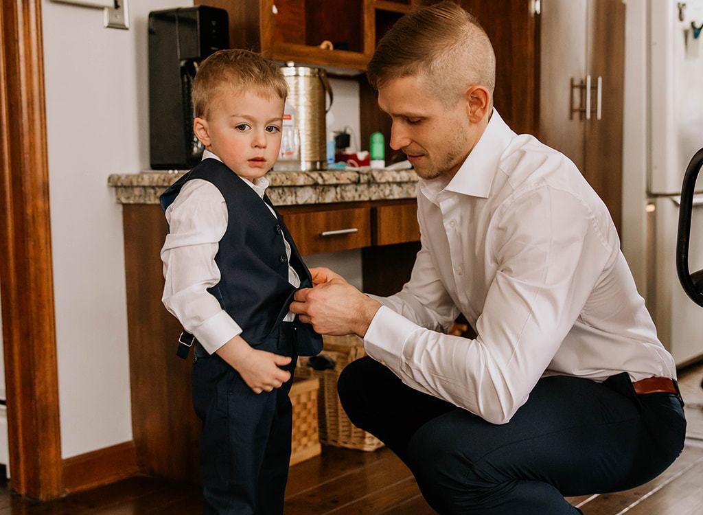 A dad in a white dress shirt and slacks bends down to help fasten his son's black suit vest. They are in a kitchen getting ready for an event.