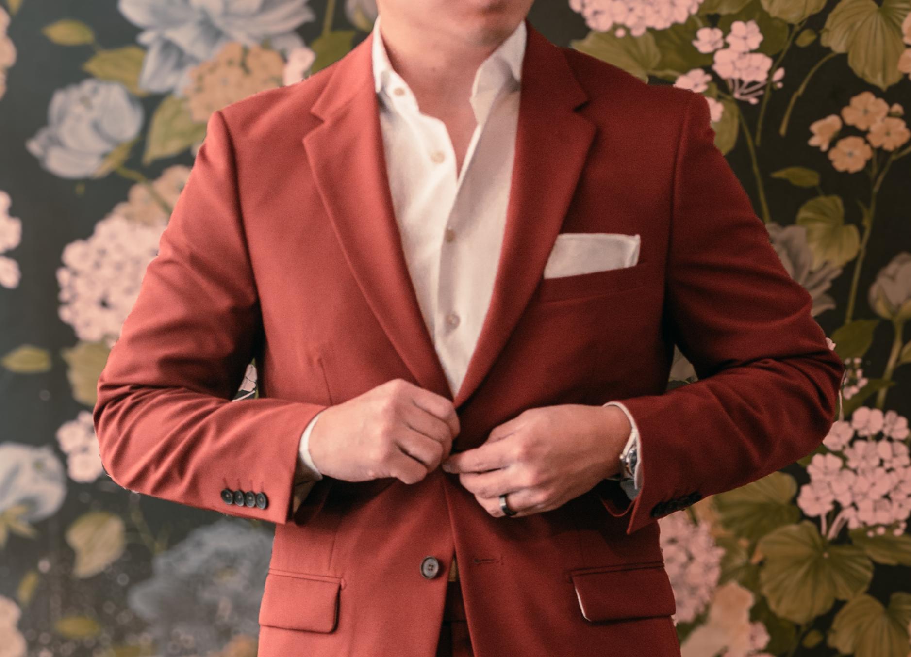 A close-up of a man's midsection as he buttons his red suit. He stands in front of floral wallpaper.