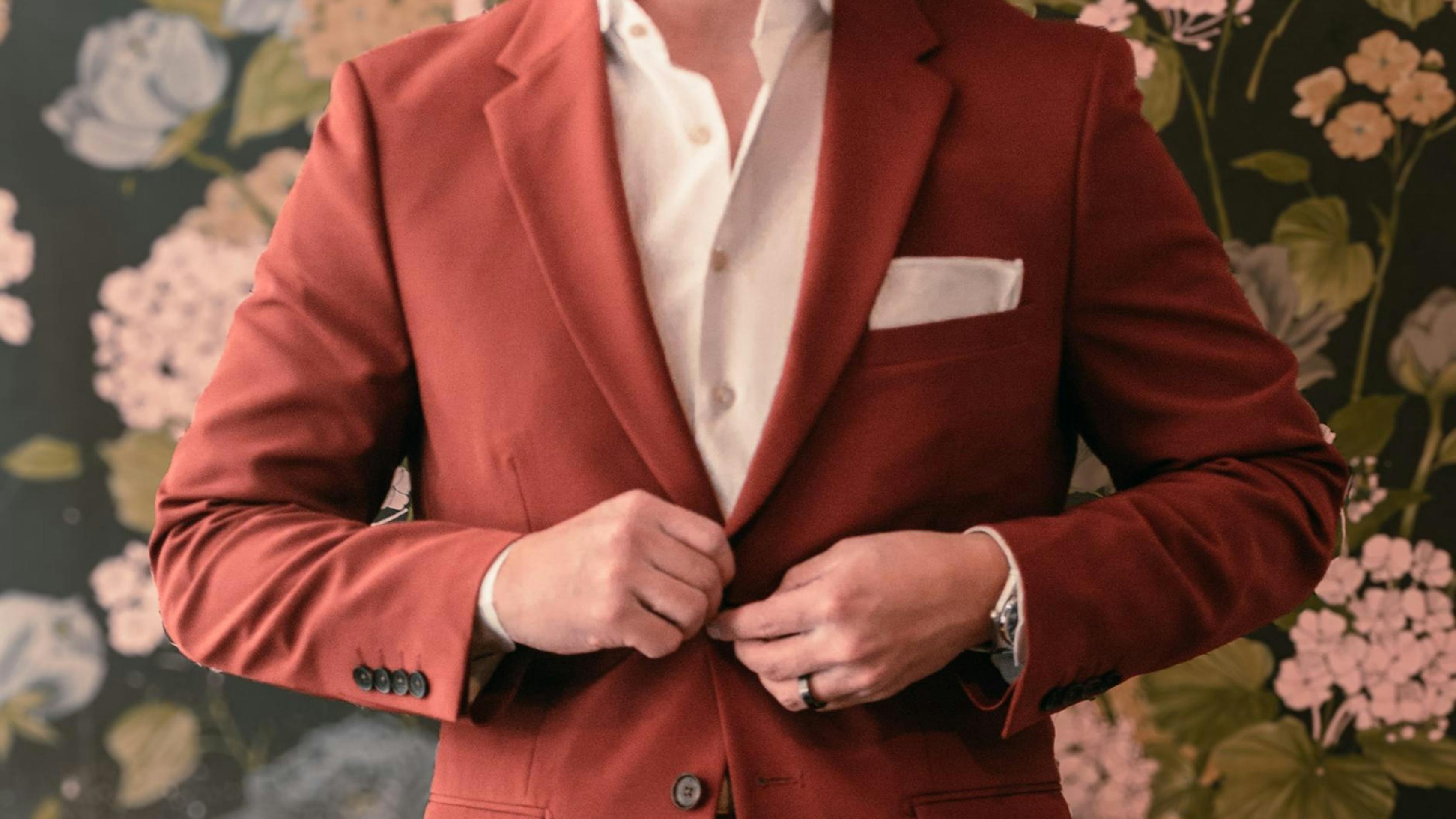 A close-up of a man's midsection as he buttons his red suit. He stands in front of floral wallpaper.