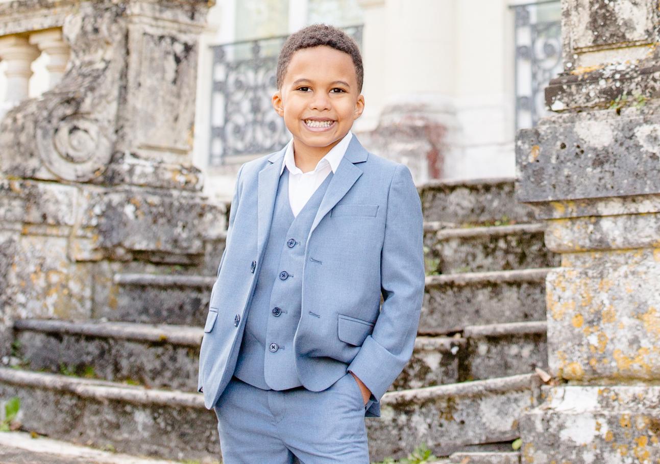A boy in a white blue suit stands in front of an old staircase leading up to a church. He smiles with his hand in his pocket.