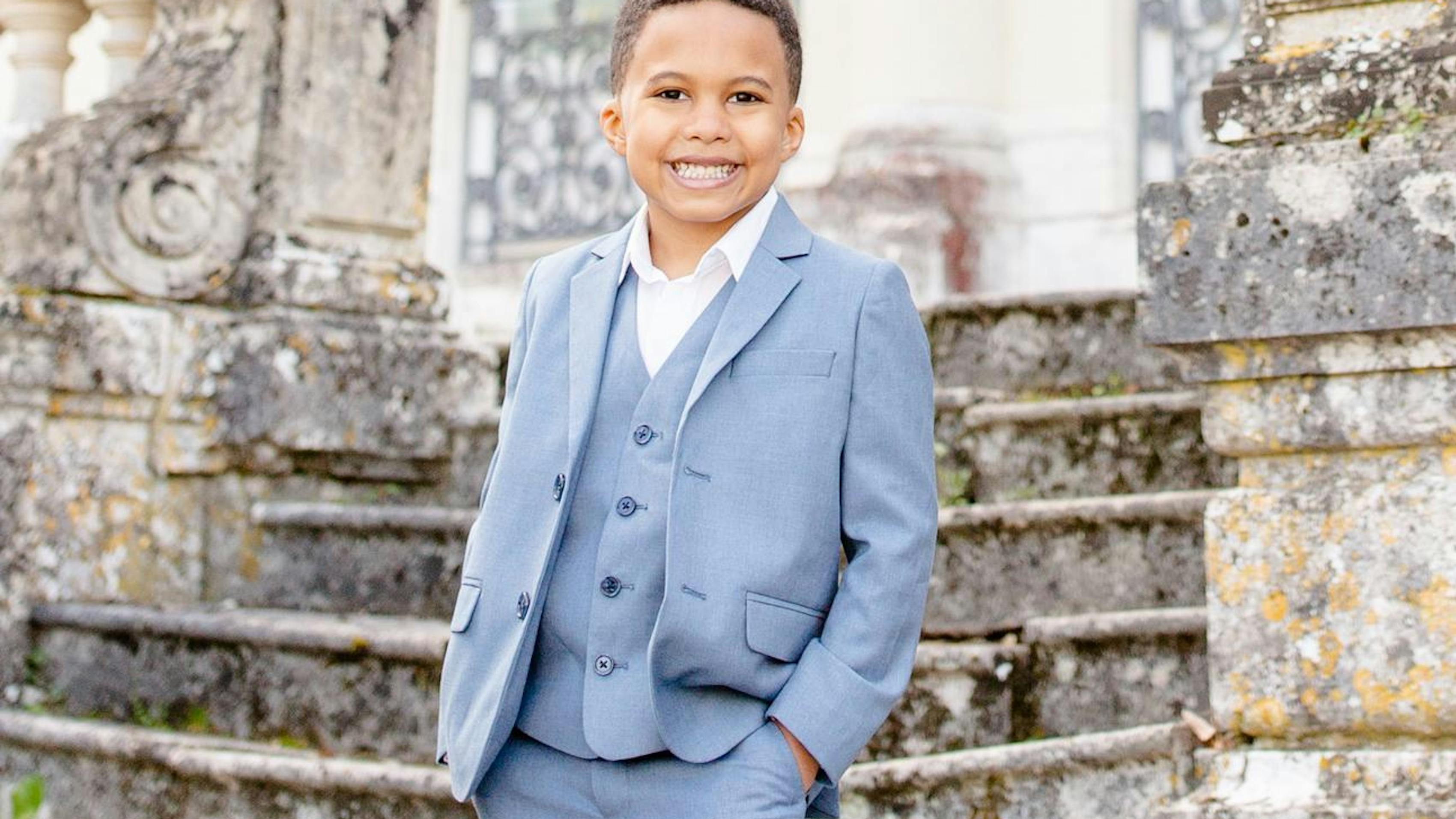 A boy in a white blue suit stands in front of an old staircase leading up to a church. He smiles with his hand in his pocket.