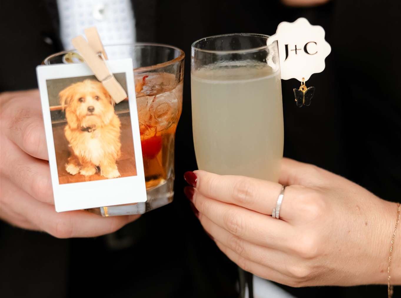 Close-up of newlyweds' hands as they clink a drink. The man is holding a mixed cocktail with a polaroid of a dog attached to it by a clip. The woman is holding a flute of champagne.