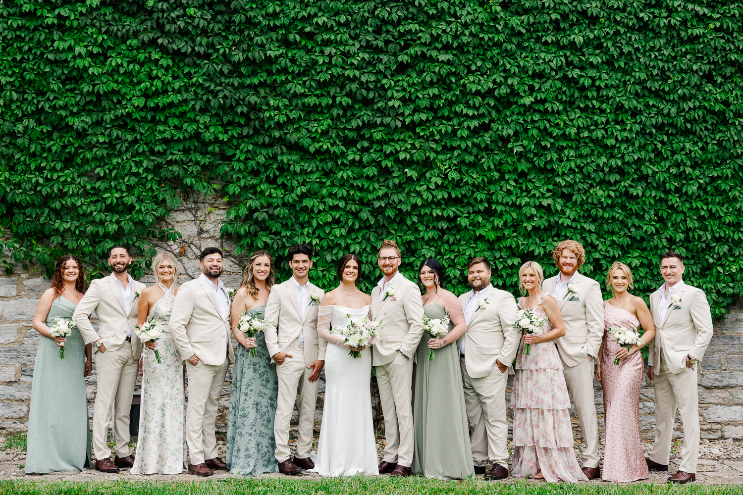 A wedding party stands around the bride and groom, all dressed in beige and pastels. They're standing against an ivy-covered wall.