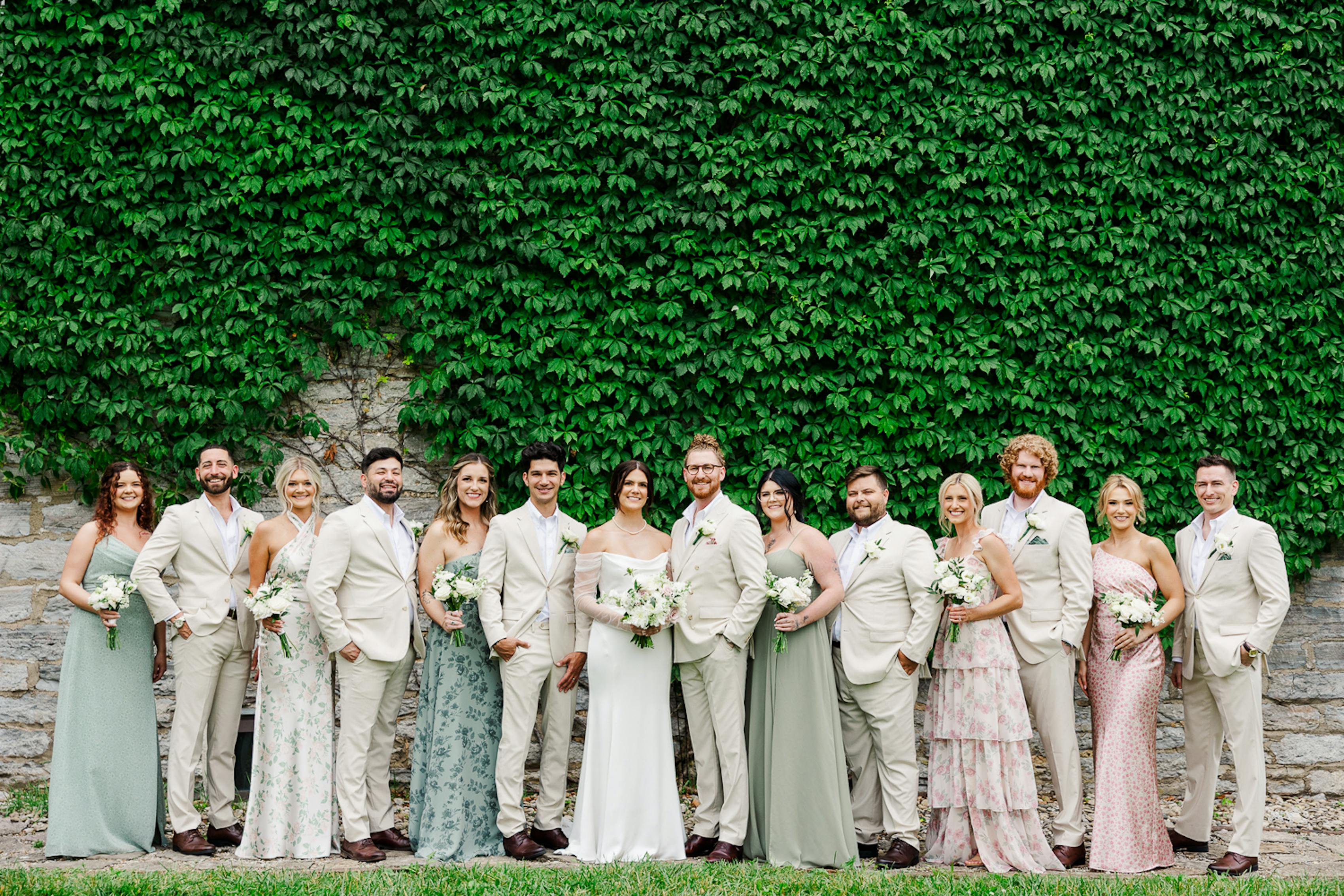 A wedding party stands around the bride and groom, all dressed in beige and pastels. They're standing against an ivy-covered wall.