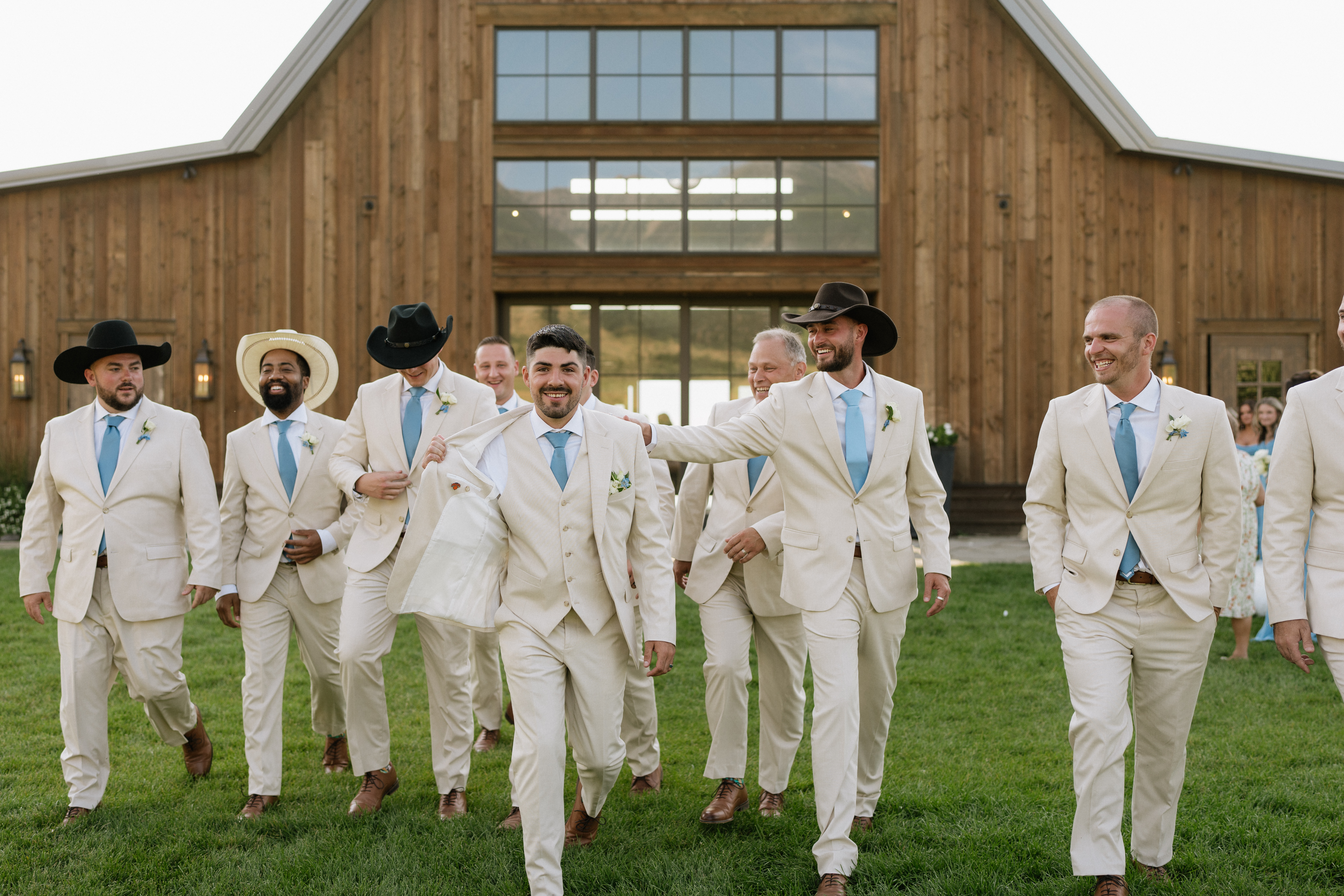 Groom's side of a wedding party, all in pastel tan suits and pastel blue ties, walking out of a barn-style wedding venye. Some of them are in cowboy hats.