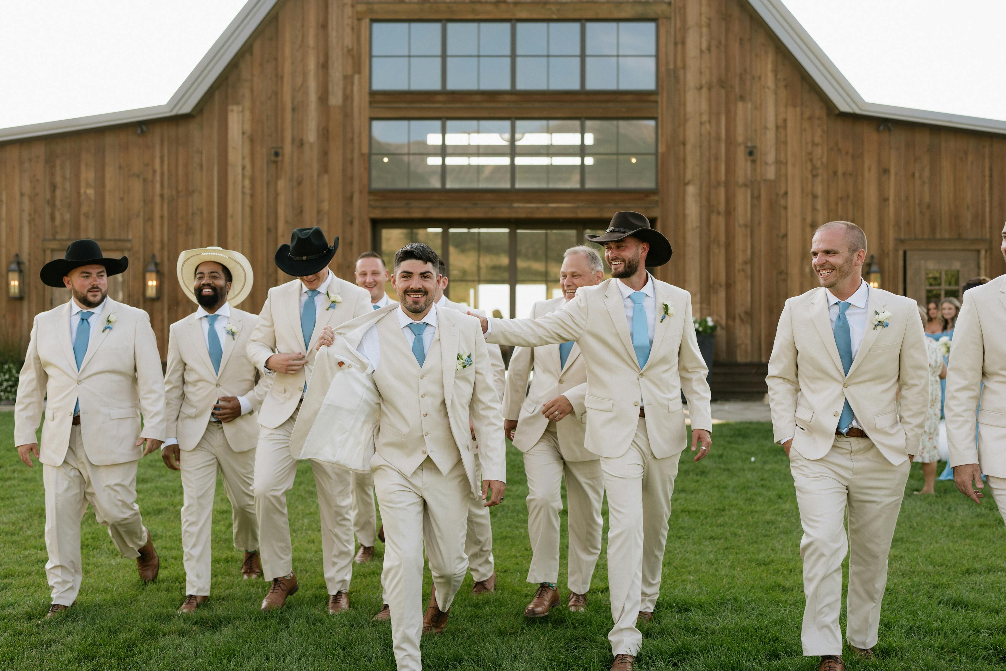 Groom's side of a wedding party, all in pastel tan suits and pastel blue ties, walking out of a barn-style wedding venye. Some of them are in cowboy hats.