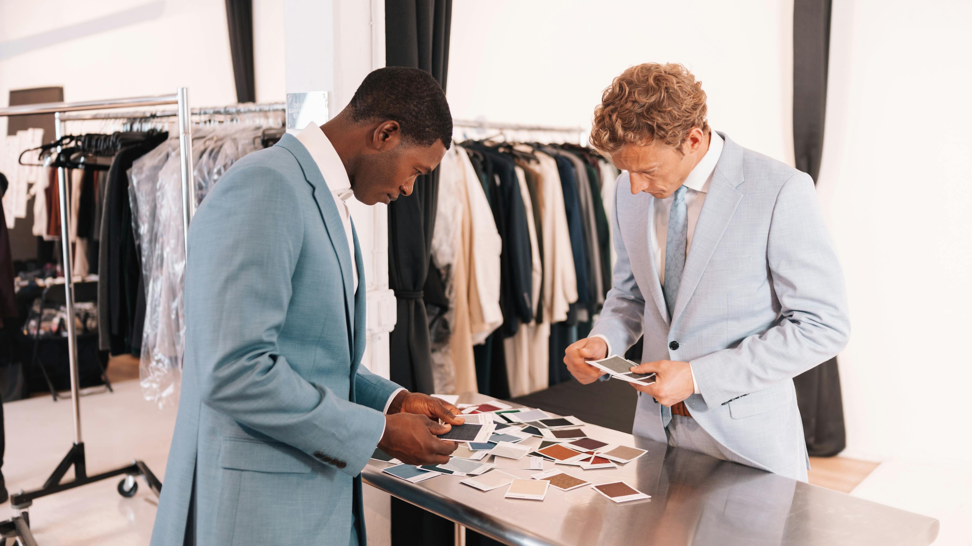 Two men in pastel blue suits at a SuitShop showroom looking through various swatches of fabric on a metal table.