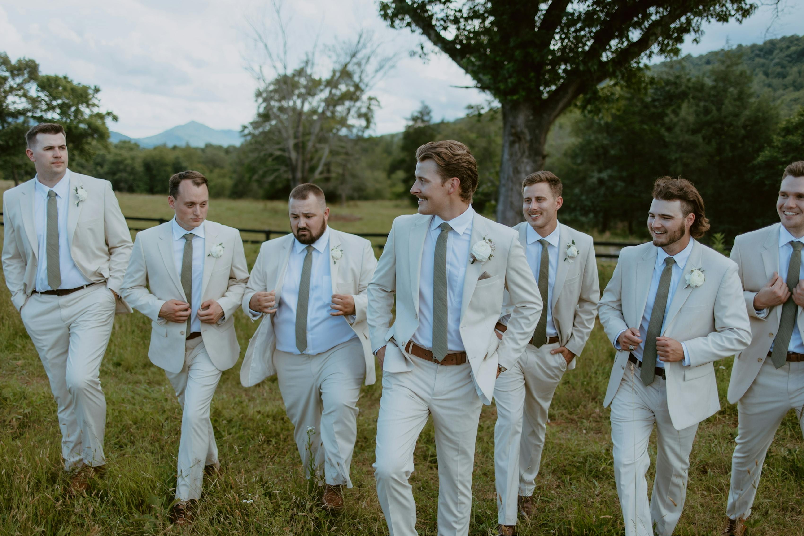 Image of the grooms' side of a wedding party walking in a field during the day. They are all dressed in tan suits.