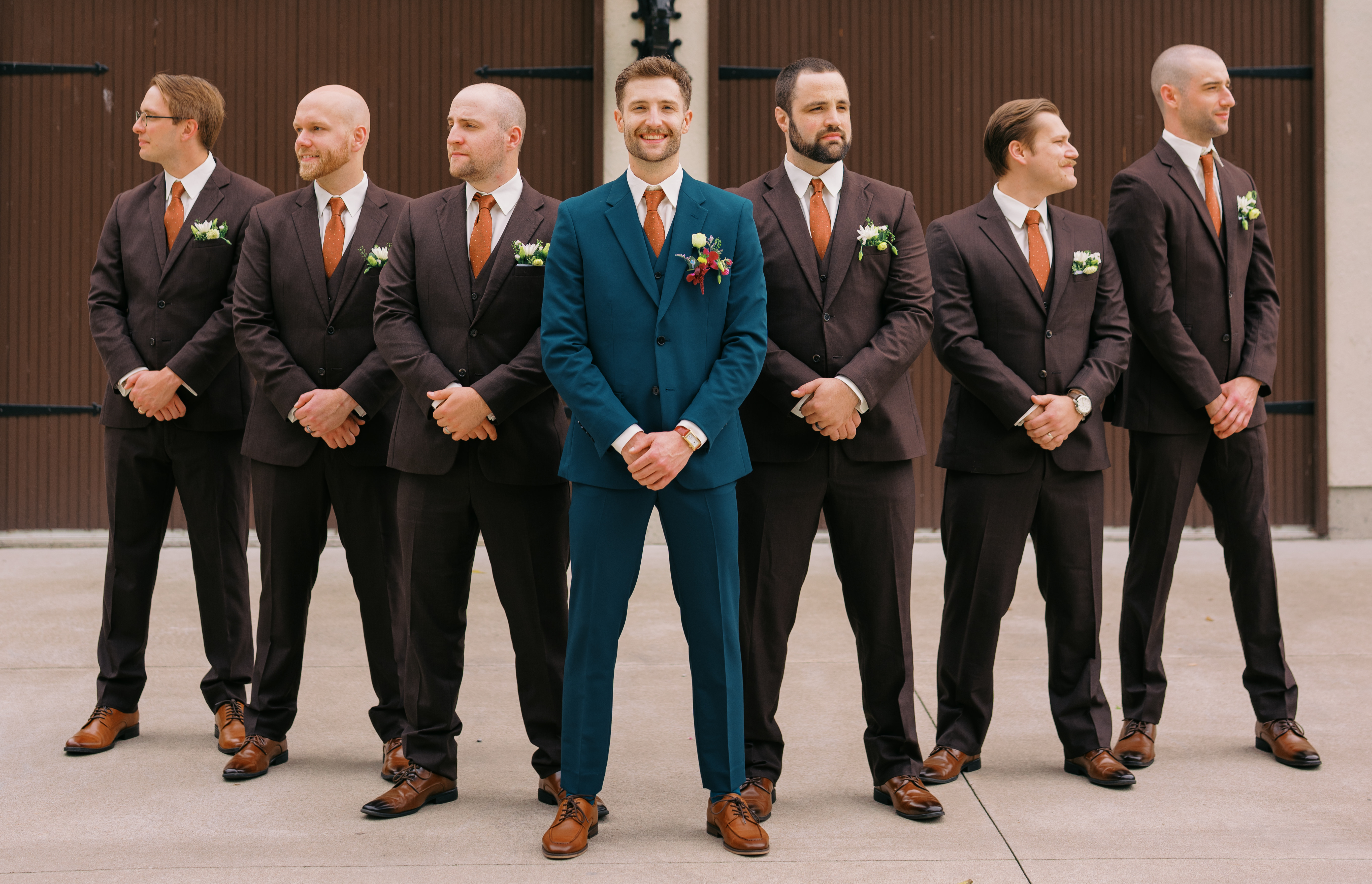 Groom's side of the wedding party posing in a driveway. The groomsmen are wearing dark brown suits while the groom is wearing a teal suit.