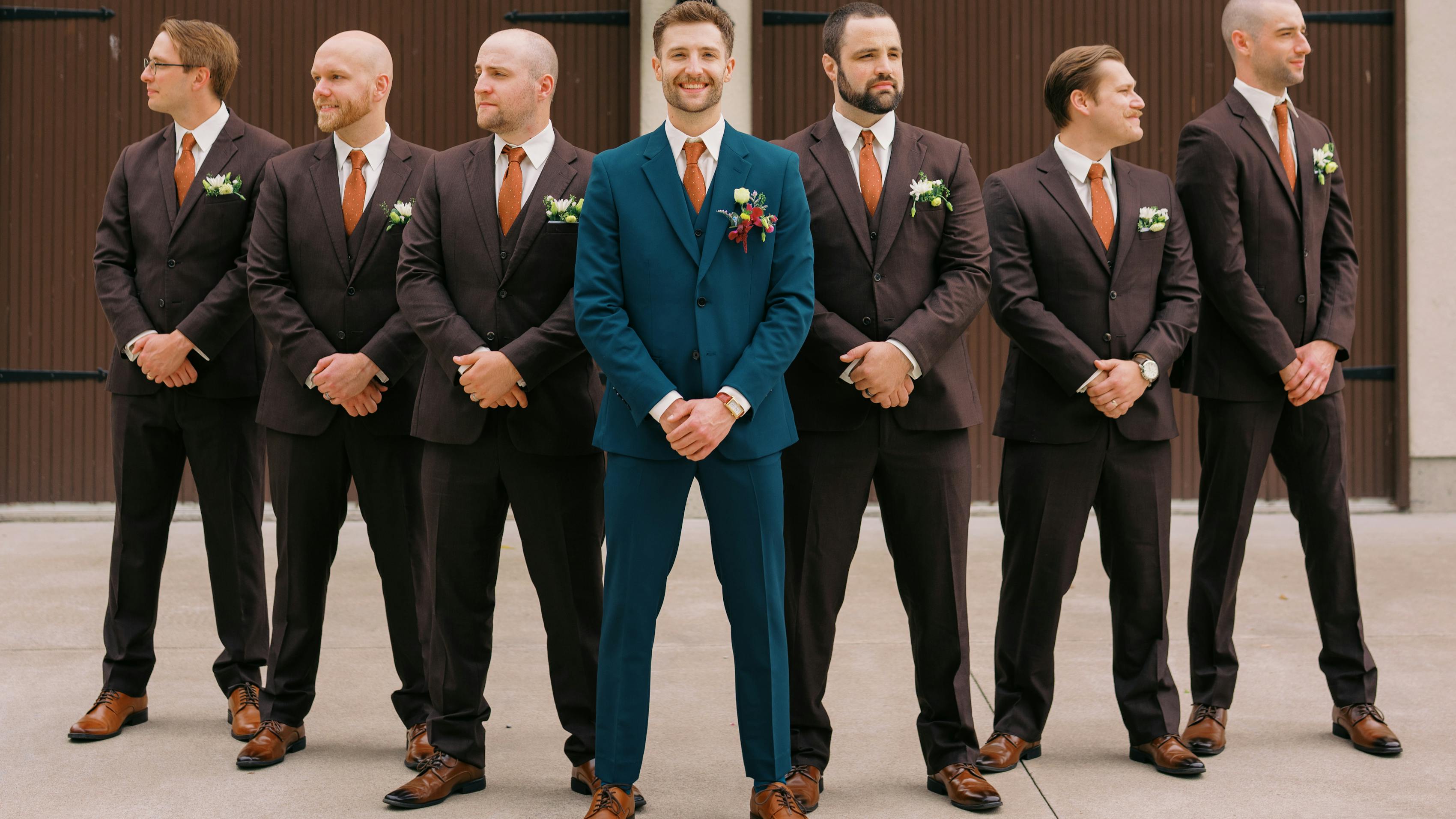 Groom's side of the wedding party posing in a driveway. The groomsmen are wearing dark brown suits while the groom is wearing a teal suit.
