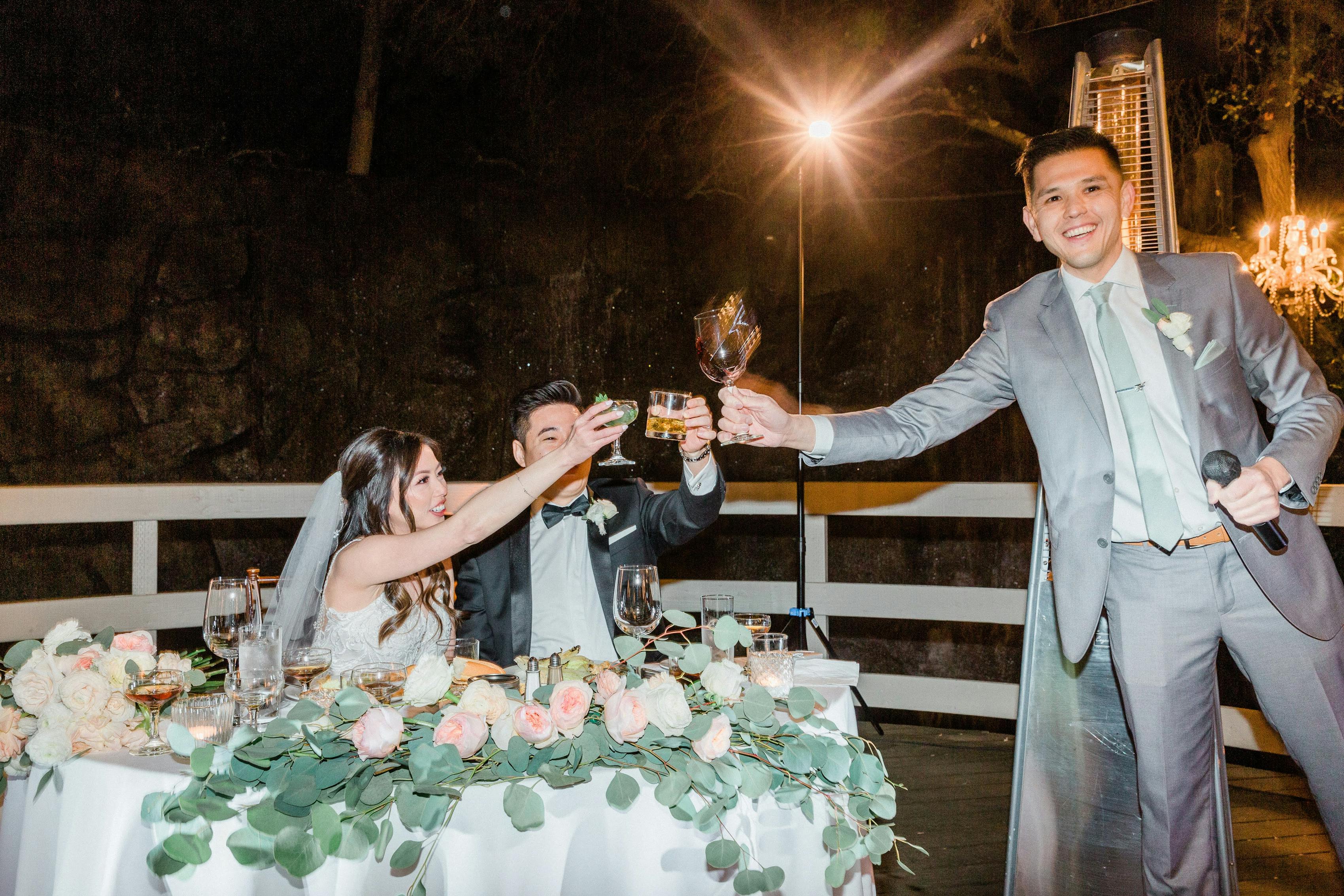 A groomsman with a microphone in his hand toasts the newlywed couple, who are sitting at a table at a wedding reception.