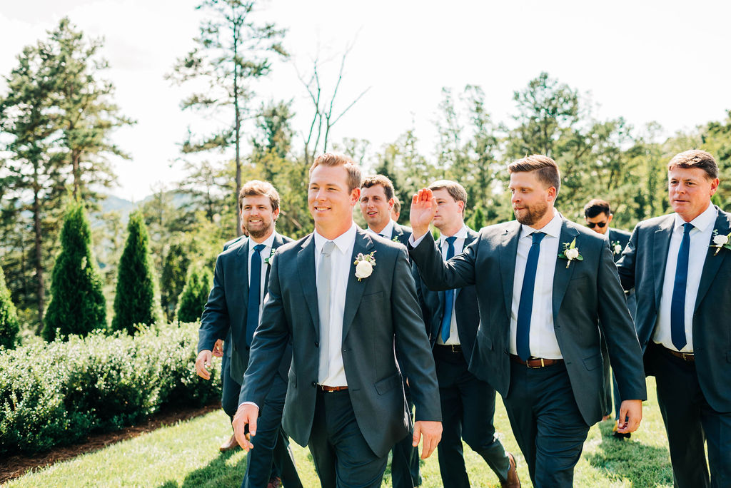 A groom walking in a field is followed by all of his groomsmen. One reaches out to pat his back.