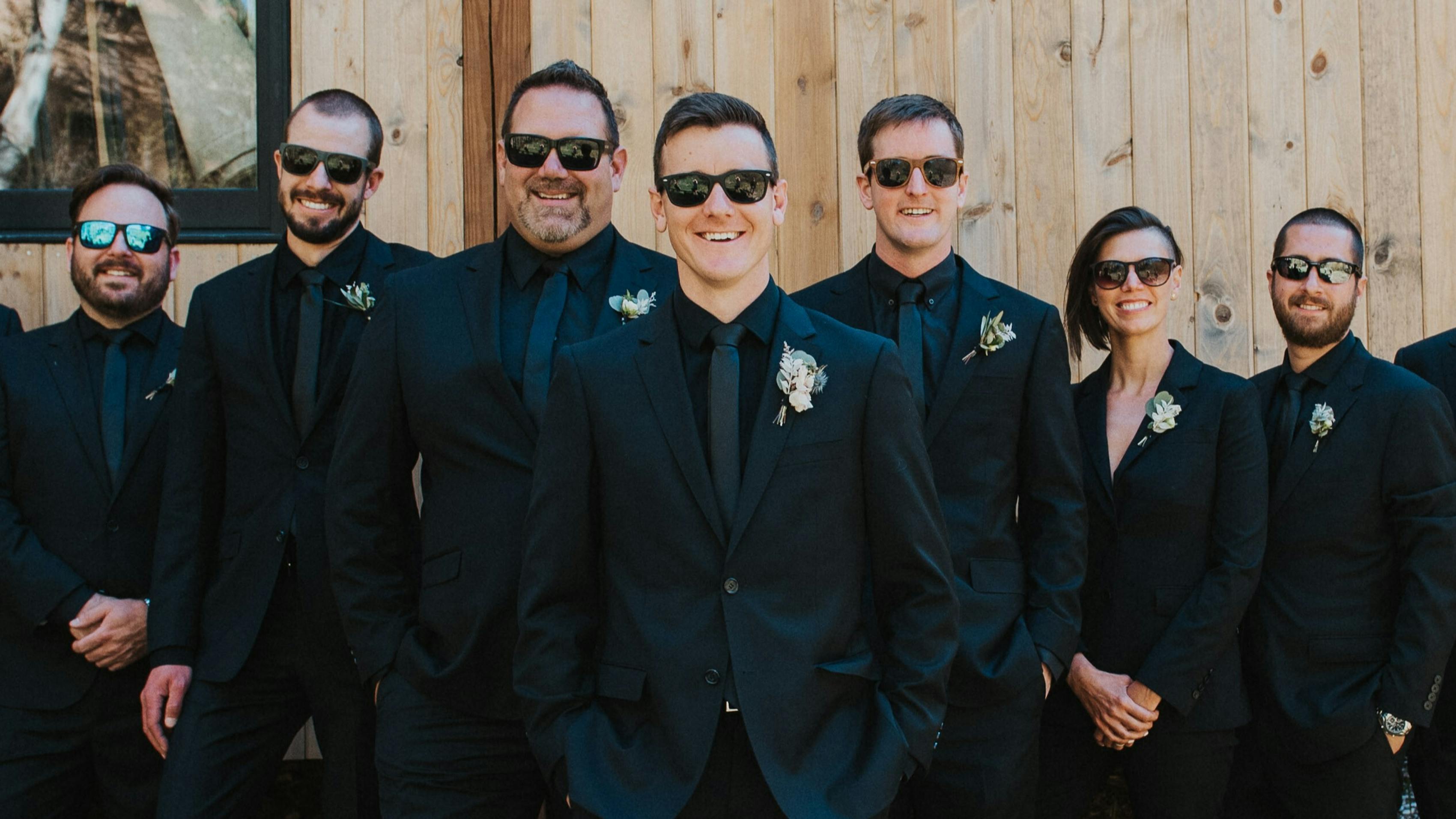 The groom's side of a bridal party, all dressed in black dress shirts and black suits. They're all wearing sunglasses and standing in front of the wood paneling of a home.