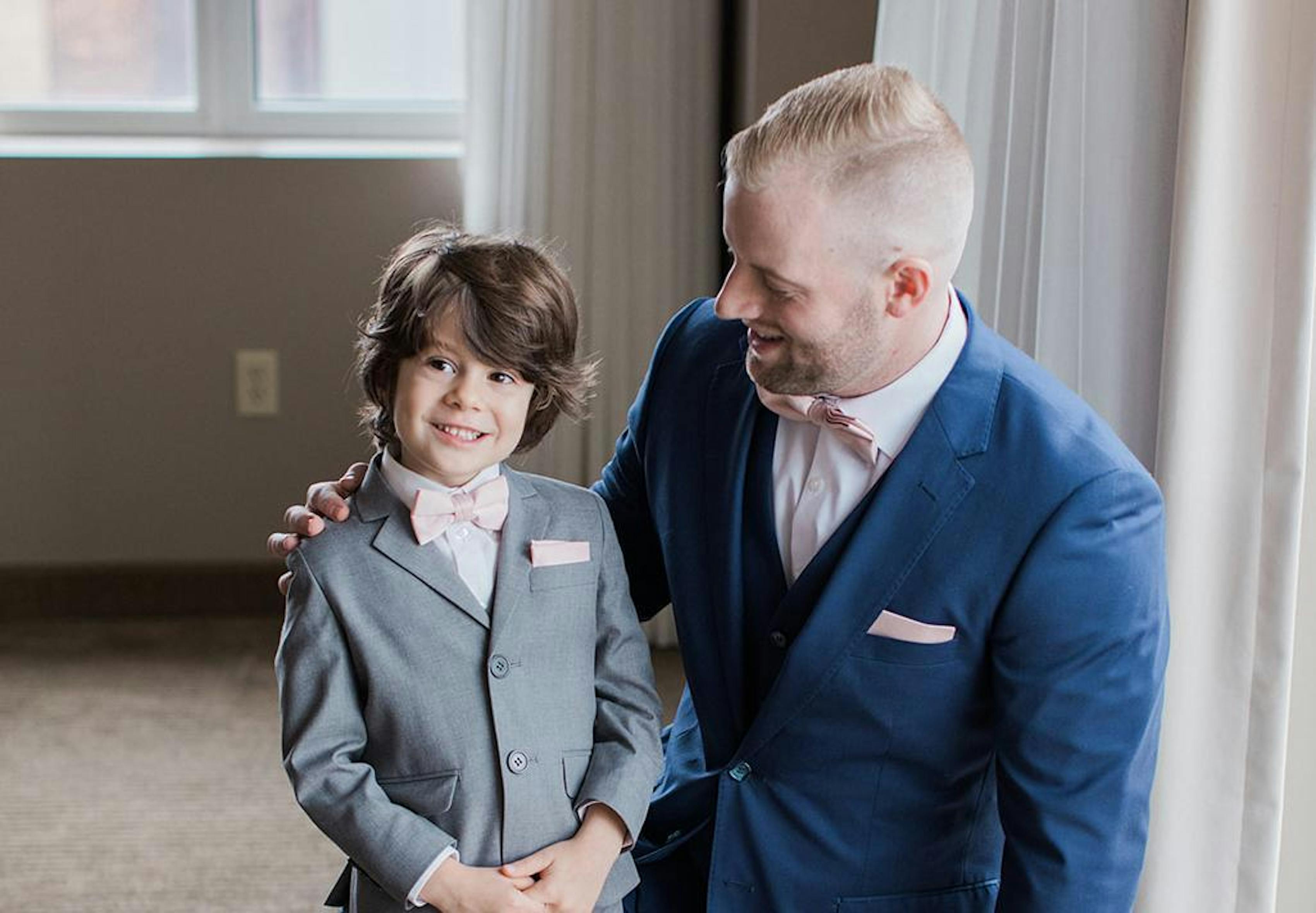 A father kneeling with his arm around his son, looking at him. The son is in a grey suit and the father is in a blue suit. They both have pink bowties and pocket squares.