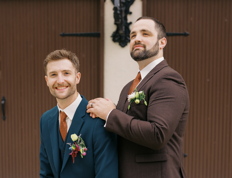 A groom and his groomsman doing a funny pose in front of a brown garage door. The groom is in a blue suit and the groomsman is in a brown suit.