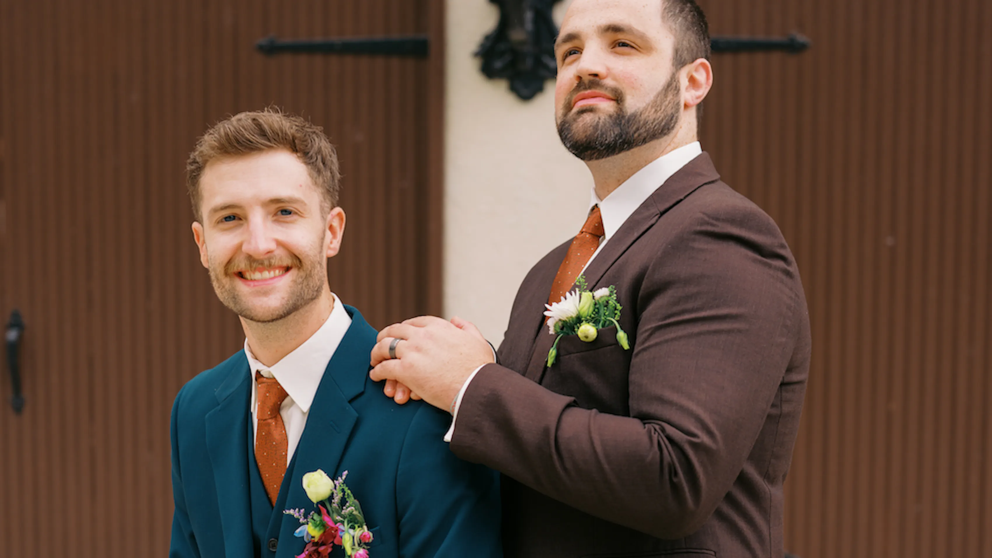 A groom and his groomsman doing a funny pose in front of a brown garage door. The groom is in a blue suit and the groomsman is in a brown suit.