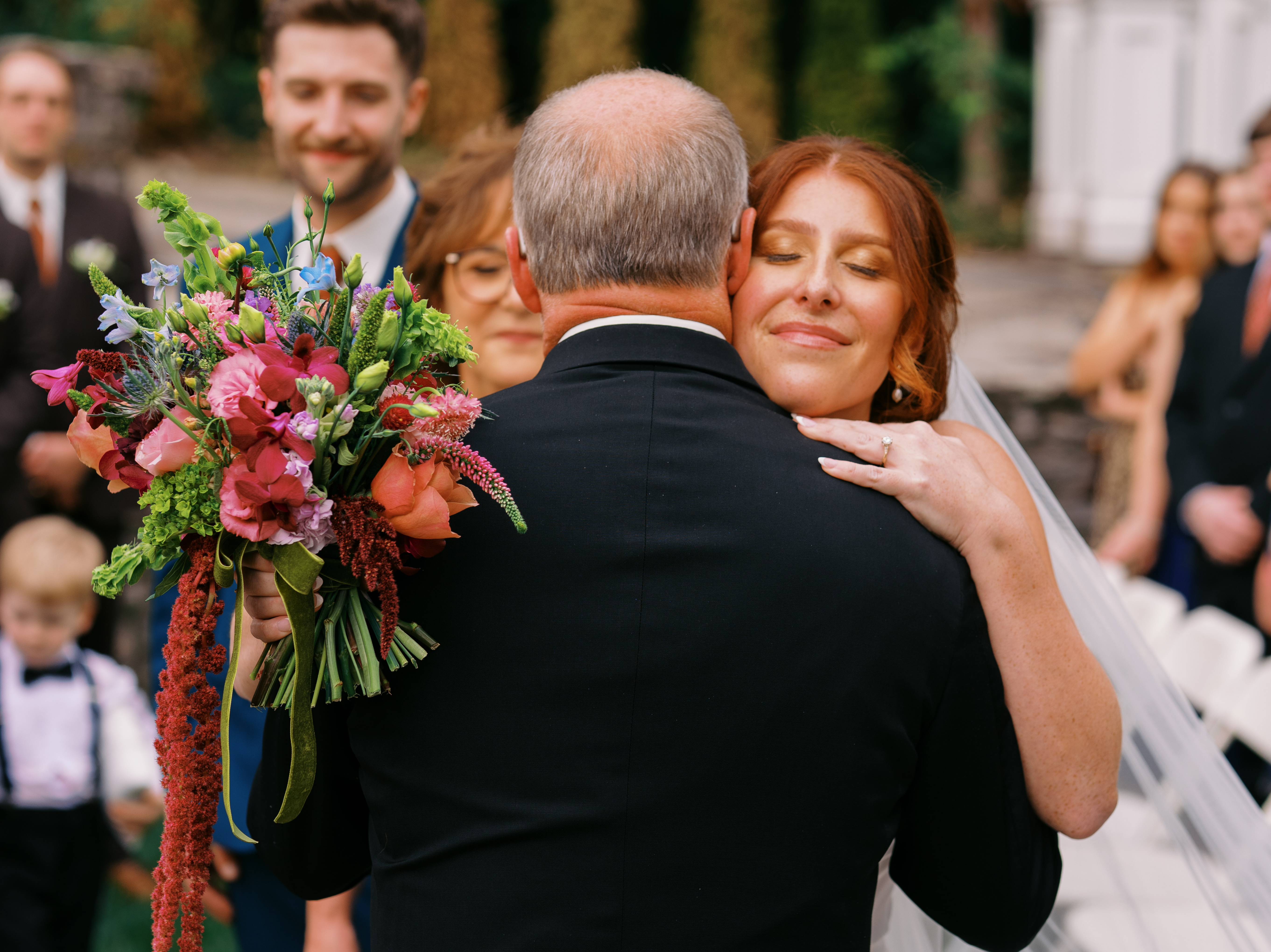 A bridge hugs her father, who is facing away from the camera, on her wedding day. She holds a bouquet in one hand.