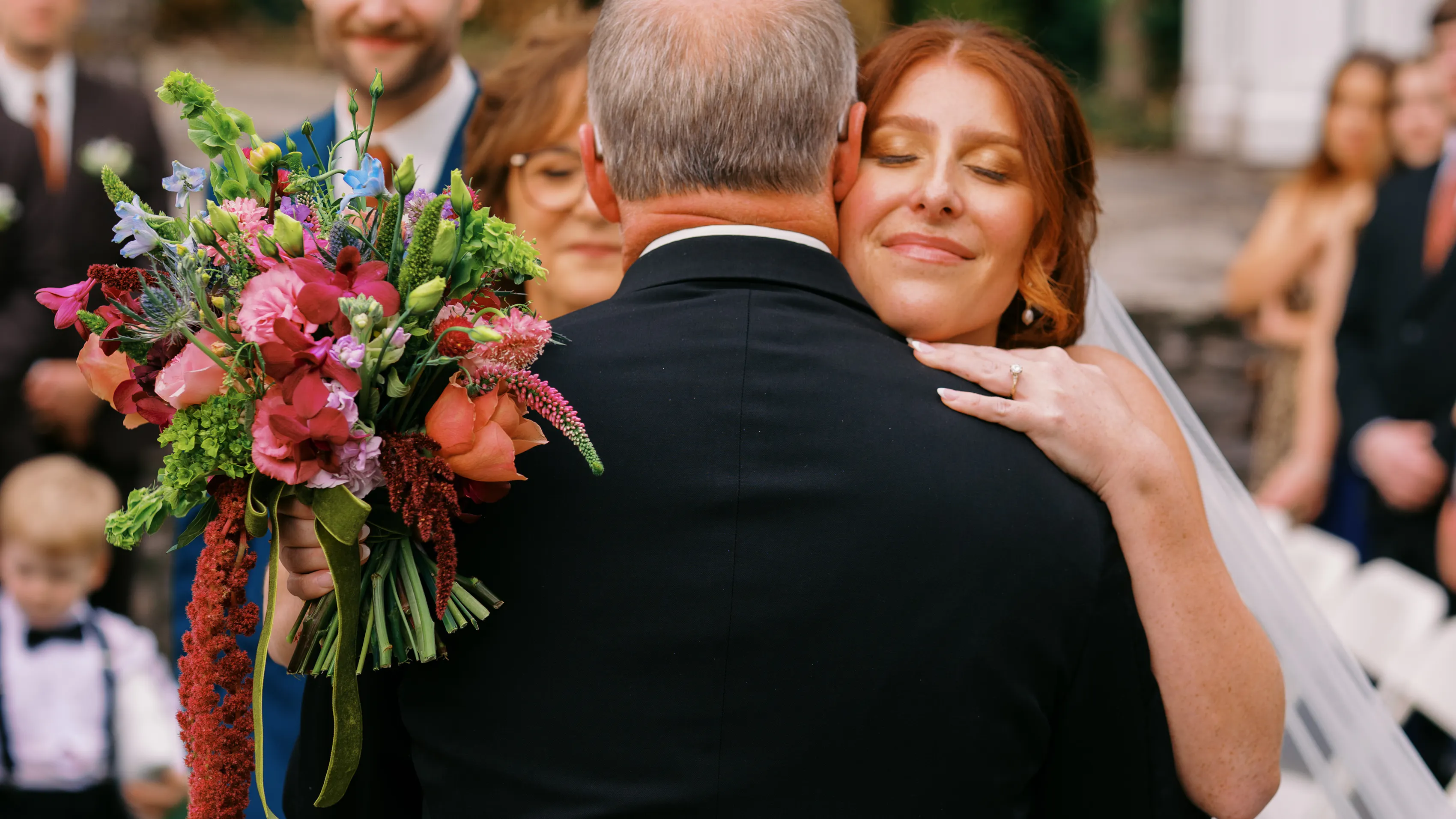 A bridge hugs her father, who is facing away from the camera, on her wedding day. She holds a bouquet in one hand.