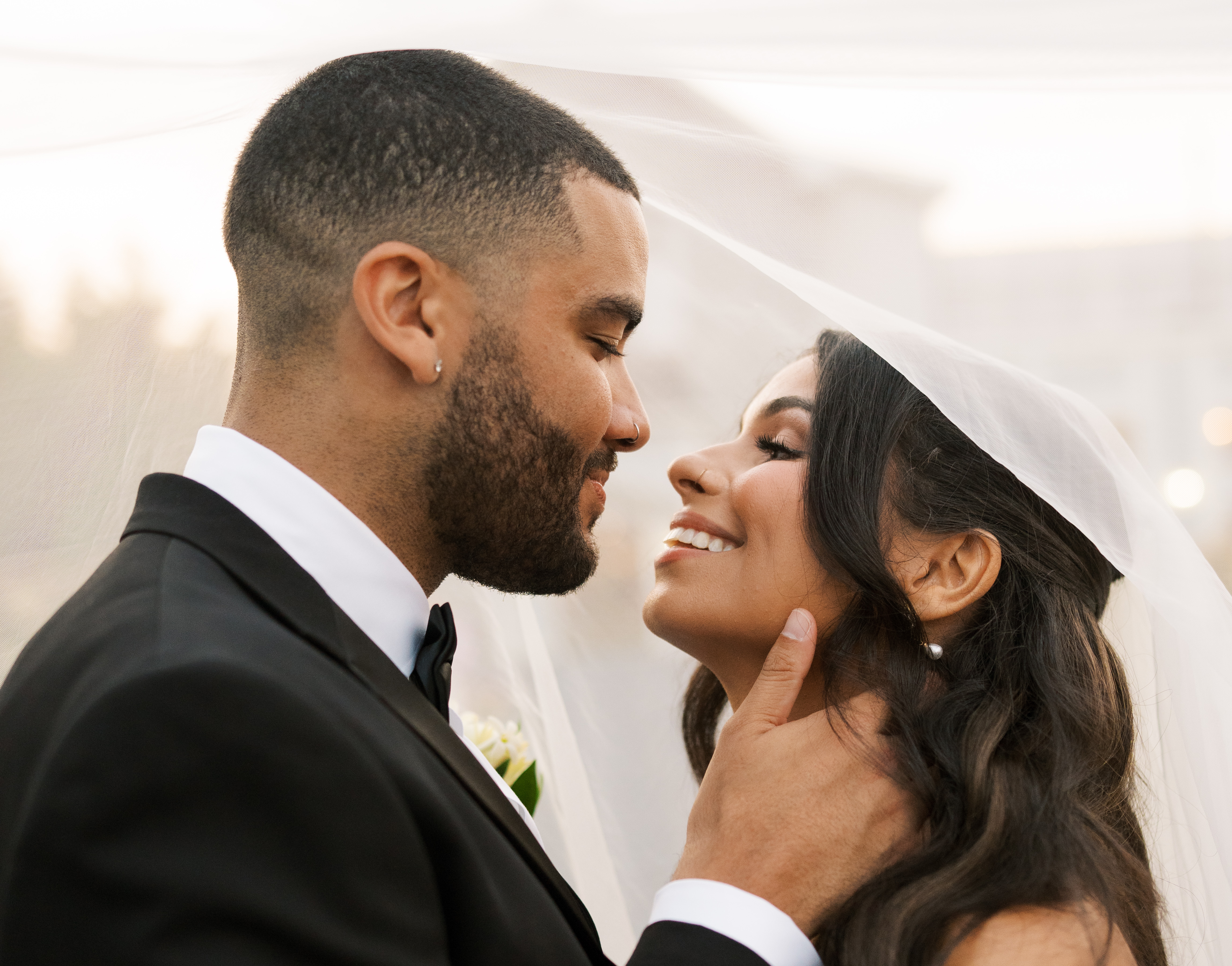 A close-up of a couple leaning in for a kiss. The man is wearing a tuxedo and is embracing the woman's neck. She is wearing a veil.