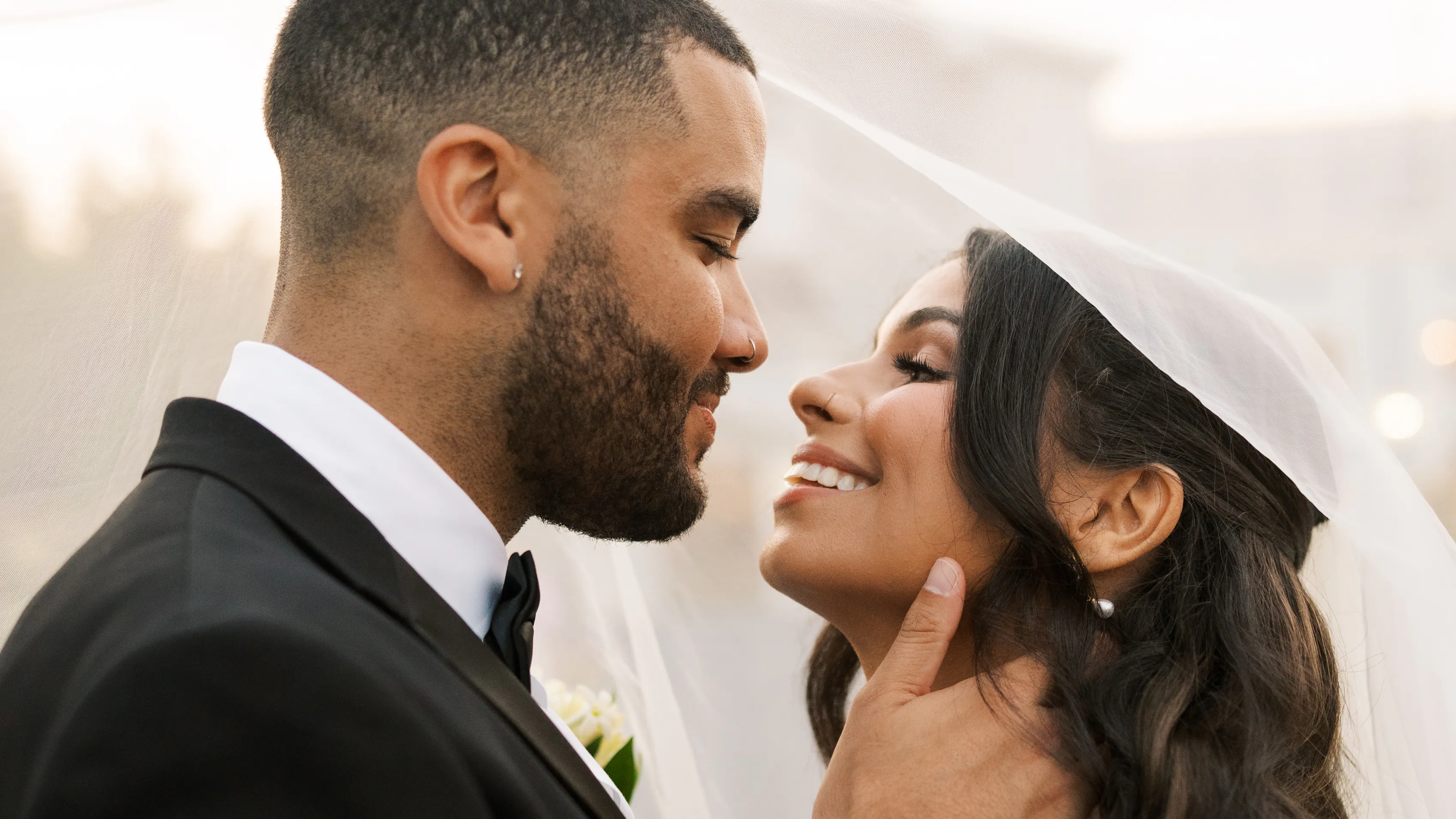 A close-up of a couple leaning in for a kiss. The man is wearing a tuxedo and is embracing the woman's neck. She is wearing a veil.
